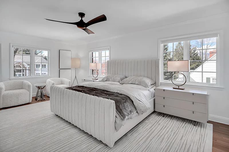 Bright modern bedroom with white vertical-paneled bed, gray striped rug, cream armchair, large windows, and ceiling fan by Cindy Montgomery.
