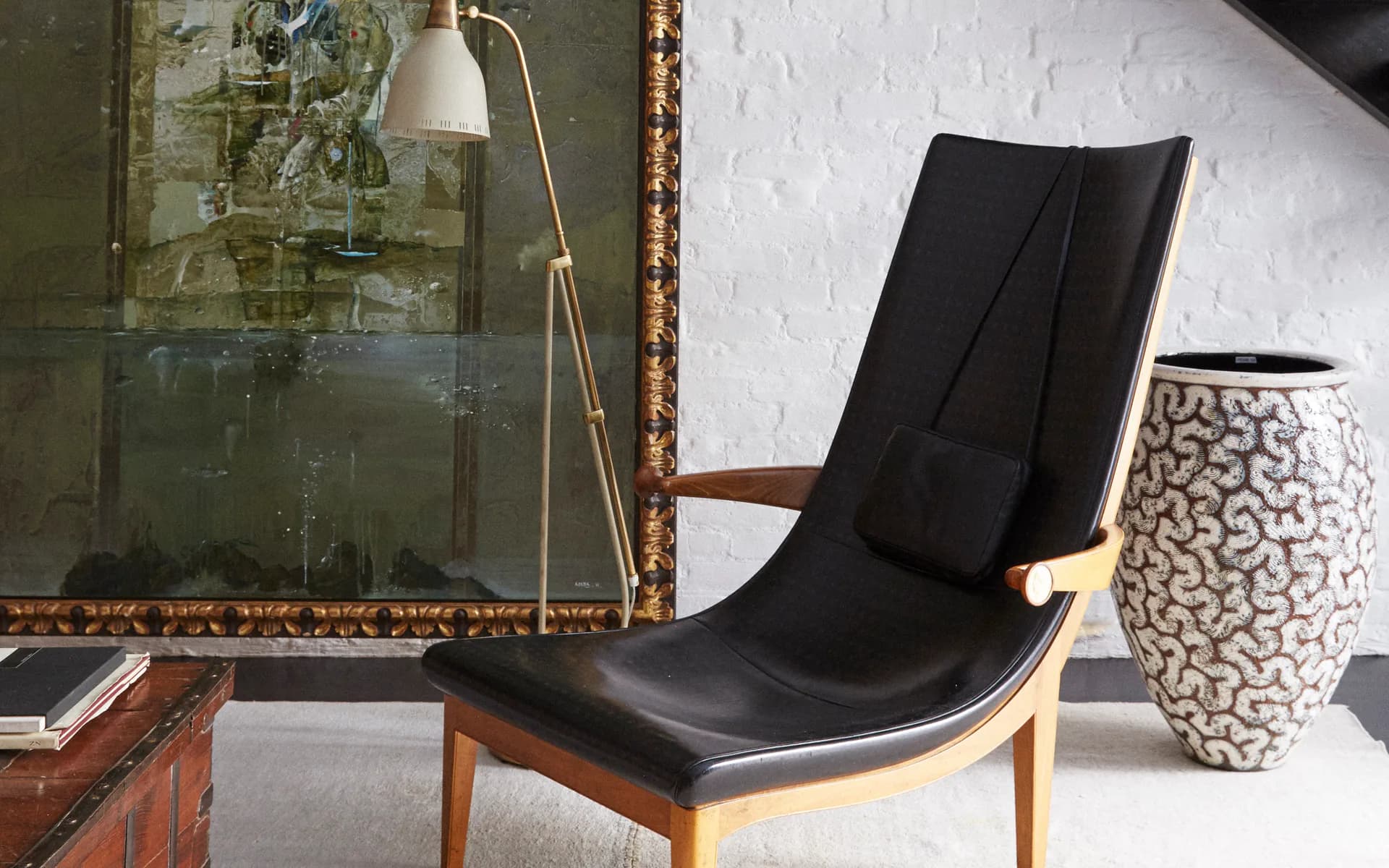 Modern interior with black leather lounge chair, wooden frame, ceramic vase, brass floor lamp, and vintage mirror on white and green walls by Claire Chung.