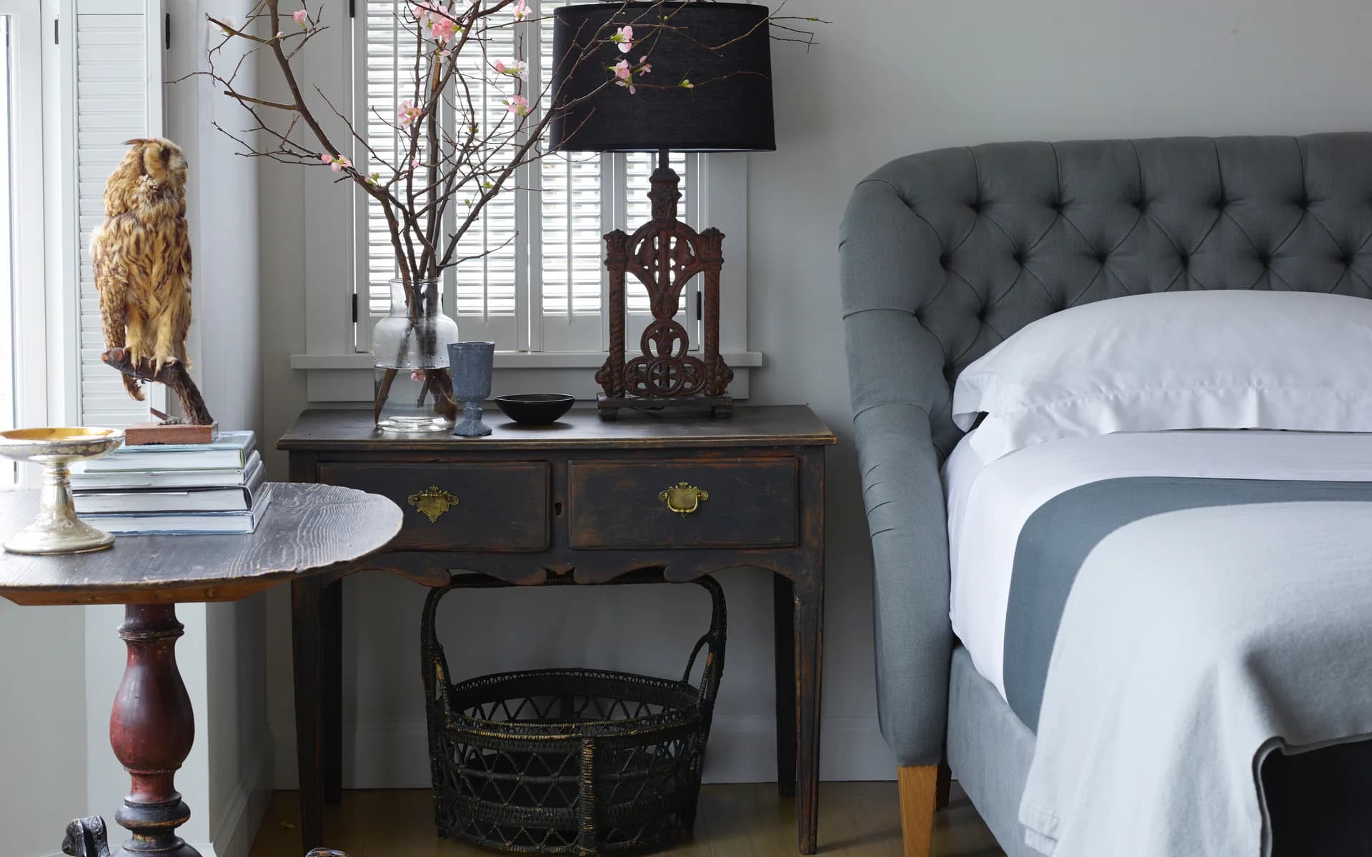 A contemporary bedroom features a tufted gray headboard, white bedding, and a dark wood desk with decorative accessories including a black lamp and botanical elements by Claire Chung.