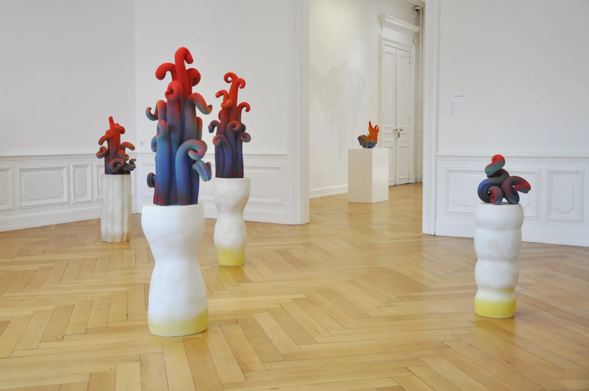 Gallery installation with sculptural figures in red and blue mounted on white pedestals across a wooden-floor exhibition space by Claire Lindner.
