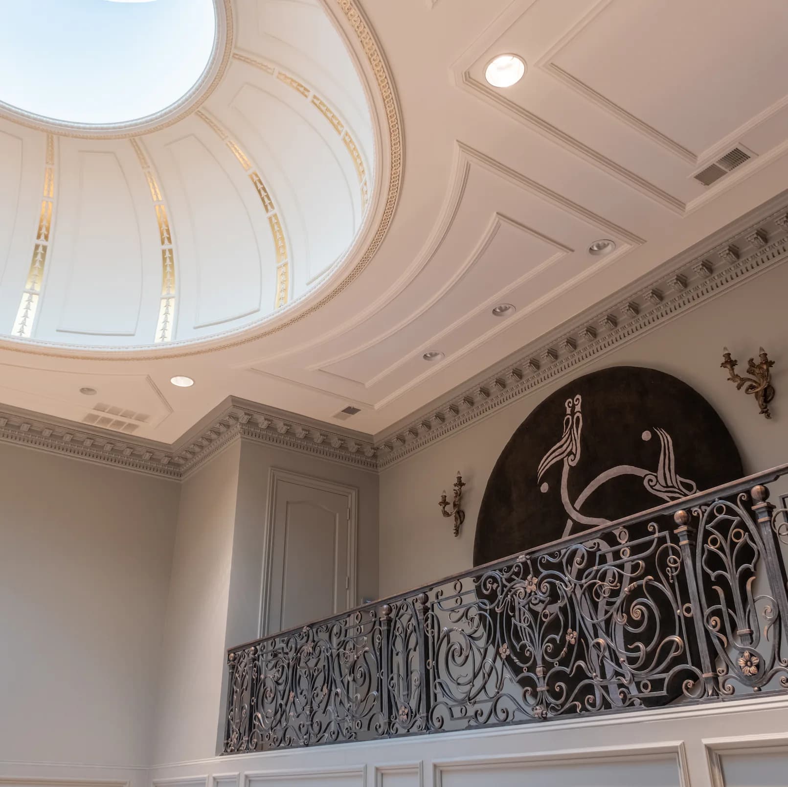 Elegant foyer with coffered ceiling, circular skylight, ornate wrought-iron balustrade, and dark accent wall featuring decorative relief by Claire McGovern.
