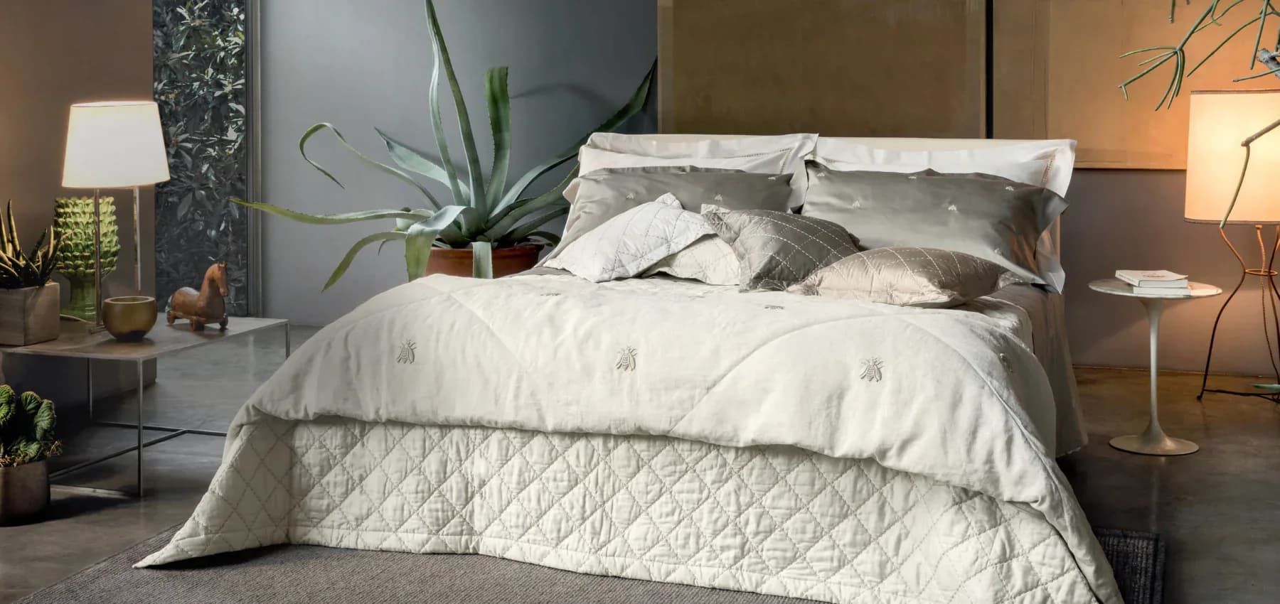 Contemporary bedroom with neutral bedding, gray upholstered headboard, warm ambient lighting, potted plants, and layered textures by Claudia Godoy.