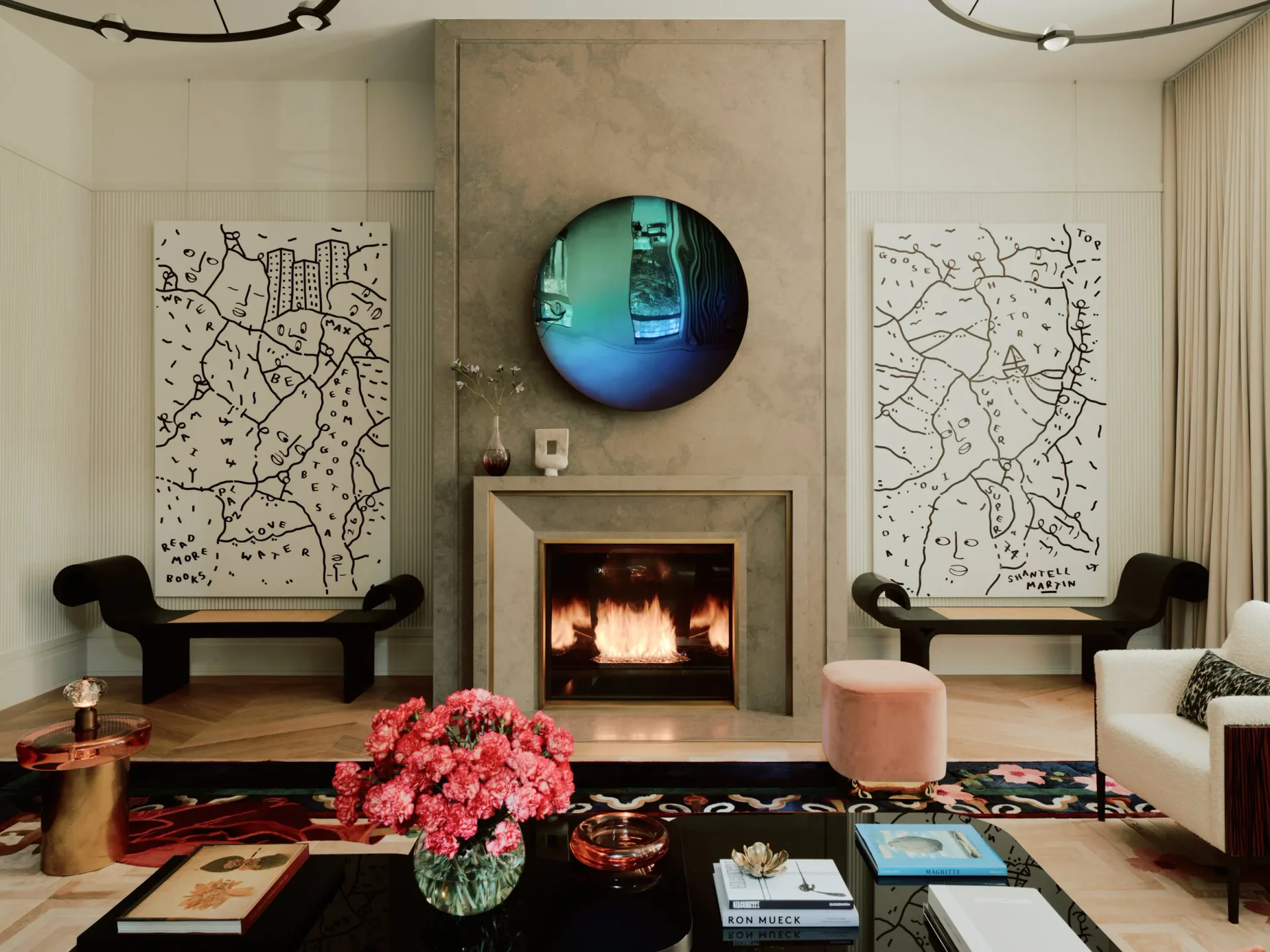 Contemporary living room with limestone fireplace, circular illuminated artwork, geometric wall maps, black seating, and pink floral arrangement by Claudia Janell.