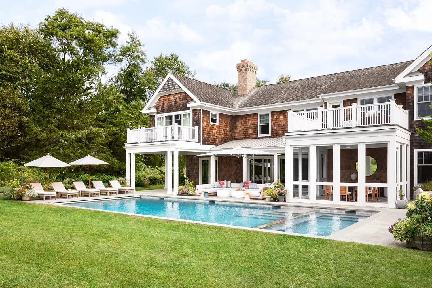 Luxurious residential backyard with brick and white colonial-style home, expansive infinity pool, manicured lawn, and covered portico overlooking grounds by Claudia Janelle.