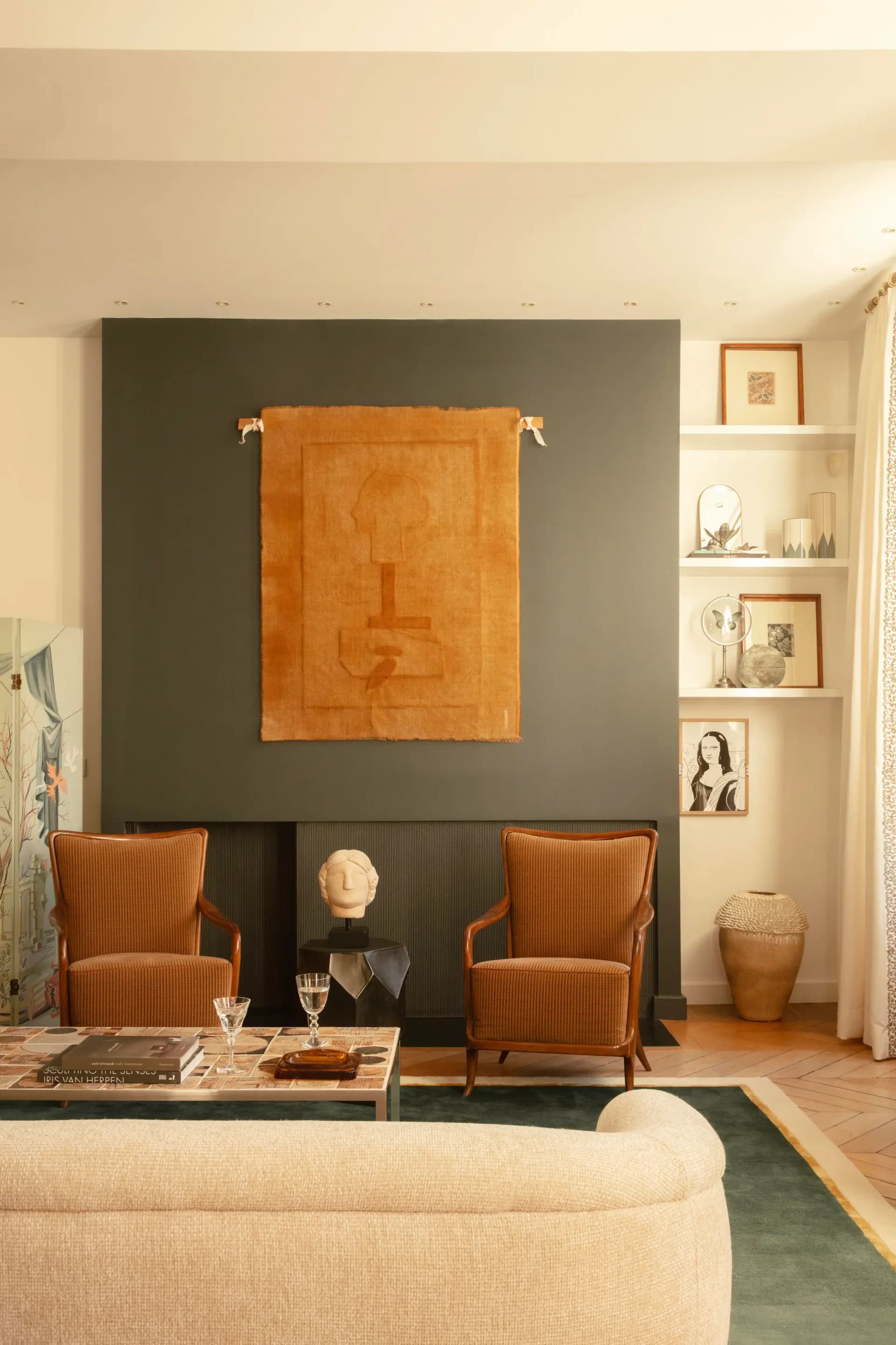 Modern living room with dark accent wall, orange abstract artwork, cognac leather chairs, built-in shelving, and neutral palette by Claudia Janelle.