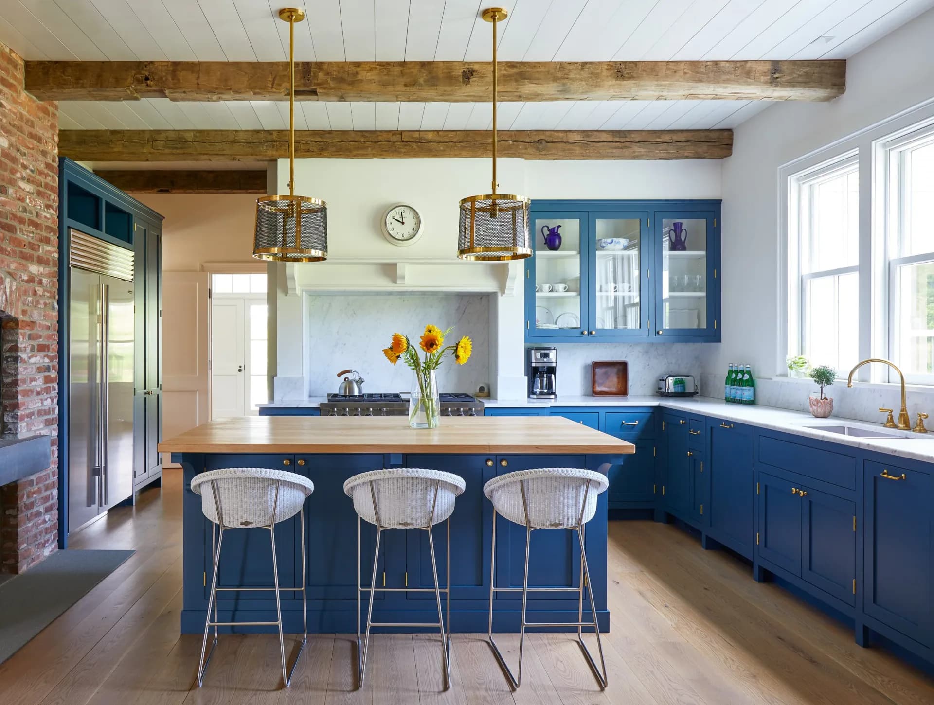 Modern kitchen with deep blue cabinetry, exposed wooden beams, brass pendant lights, white subway tile, and blue bar stools by Claudia Kalur.