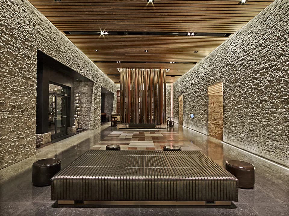 Luxurious bedroom with textured stone walls, wooden slat ceiling, geometric patterned floor, and a striking ribbed platform bed by Clodagh.