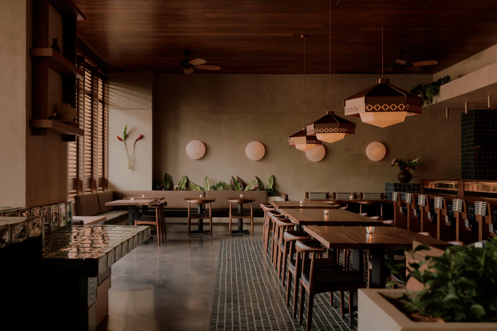 Modern restaurant interior with warm wood tones, pendant lighting, dining tables, geometric floor patterns, and potted plants throughout, designed by Colleen Smith.