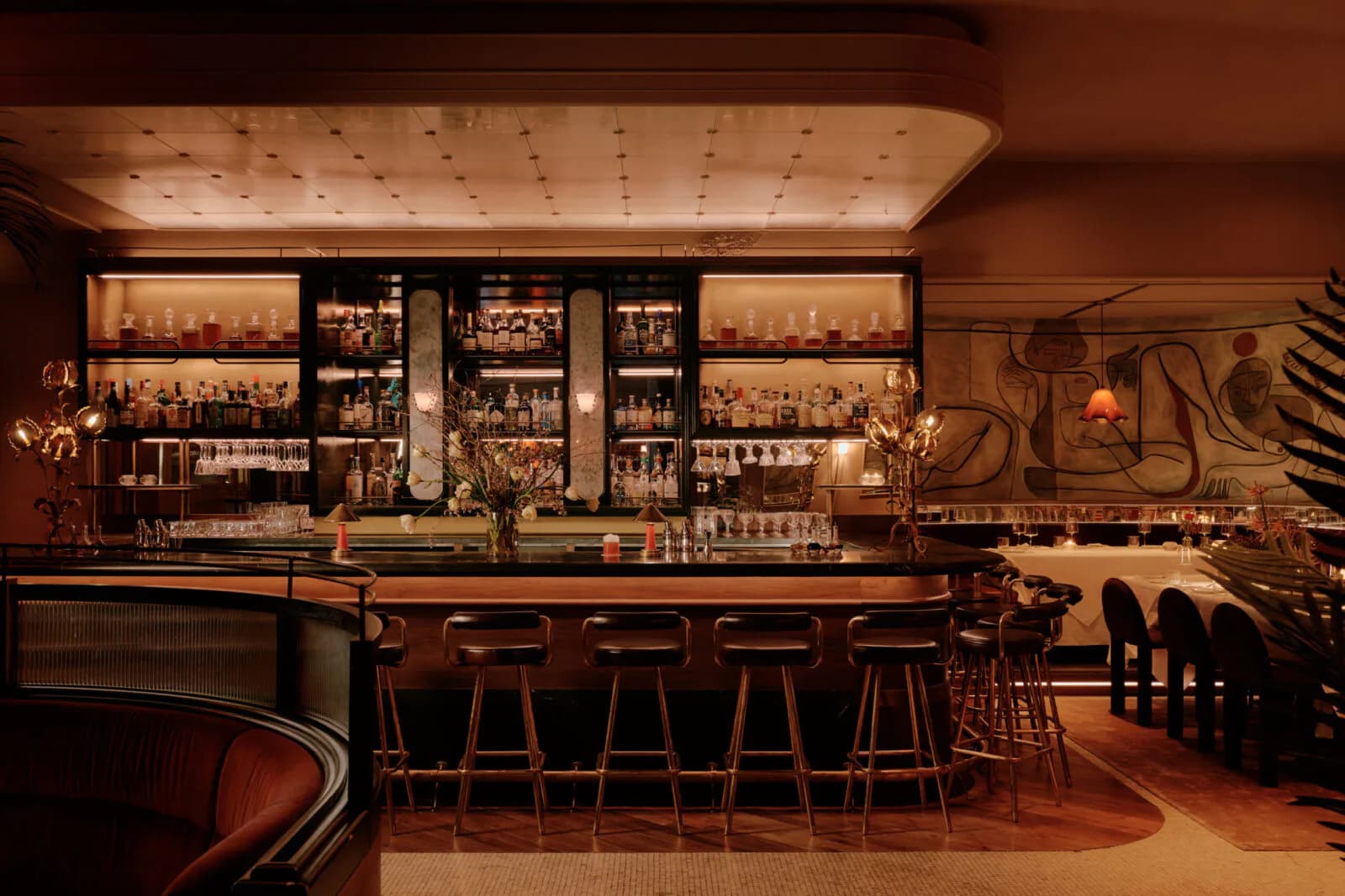 Warm-toned luxury bar interior featuring a mahogany counter, backlit shelving, brass accents, and rich jewel-toned upholstery by Colleen Smith.