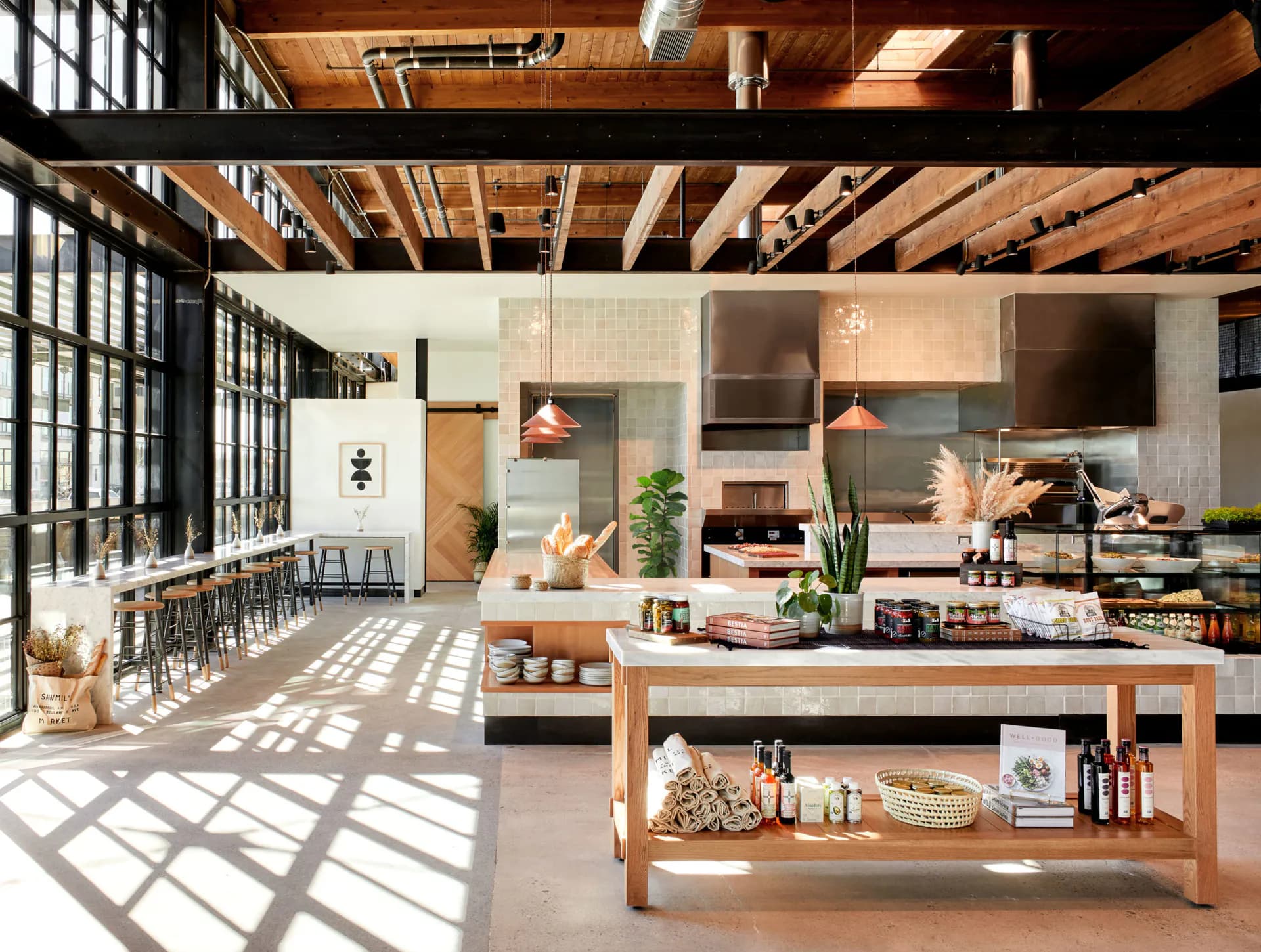 Spacious open-plan kitchen and dining area with exposed wooden ceiling beams, concrete walls, natural wood cabinetry, and large gridded windows flooding the space with light, by Colleen Smith.