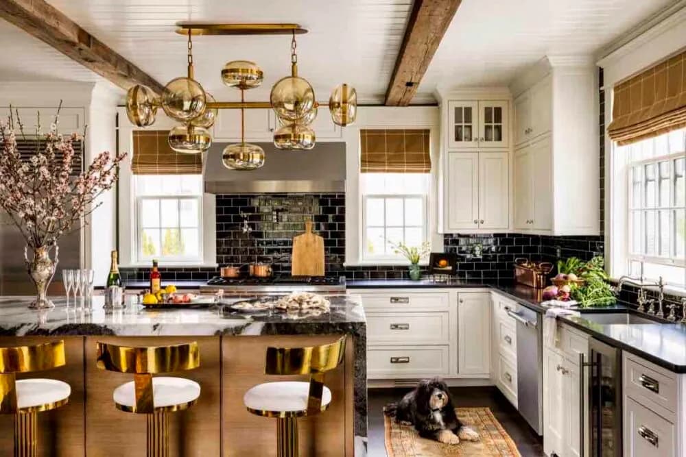 Modern kitchen with white cabinetry, black subway tile backsplash, brass pendant lights, and a wood-topped island with brass-and-cream stools by Corey Damen Jenkins.