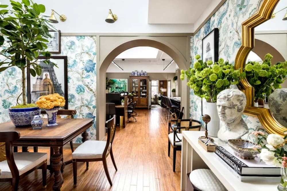 Elegant foyer with arched doorway, blue floral wallpaper, wooden floors, decorative mirrors, classical bust, and fresh greenery throughout by Corey Damen Jenkins.