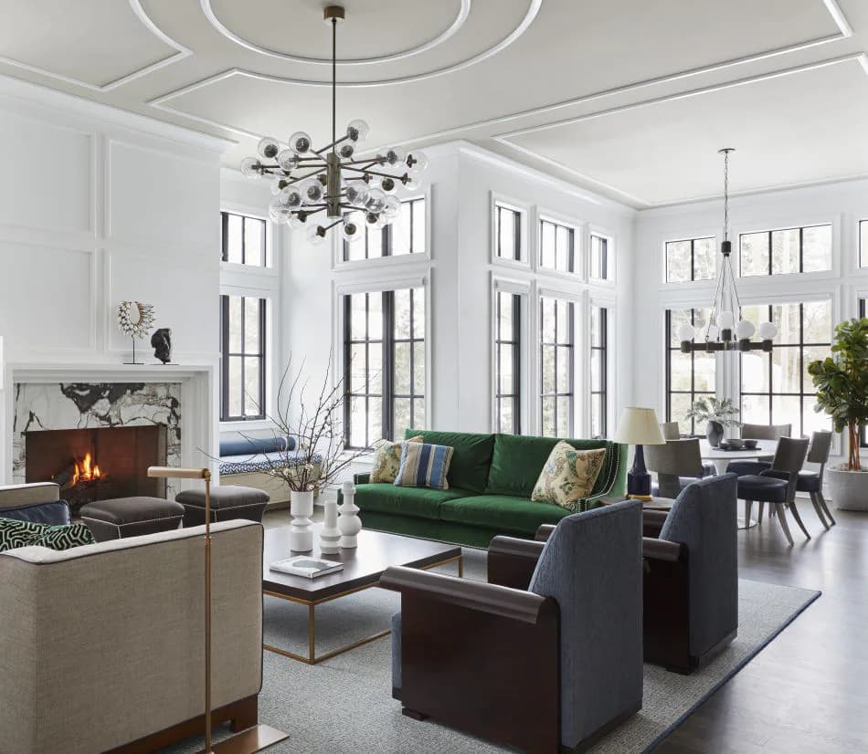Luxurious open-plan living room with high ceilings, fireplace, green velvet sofa, dark furniture, and multiple tall windows, designed by Corey Damen Jenkins.