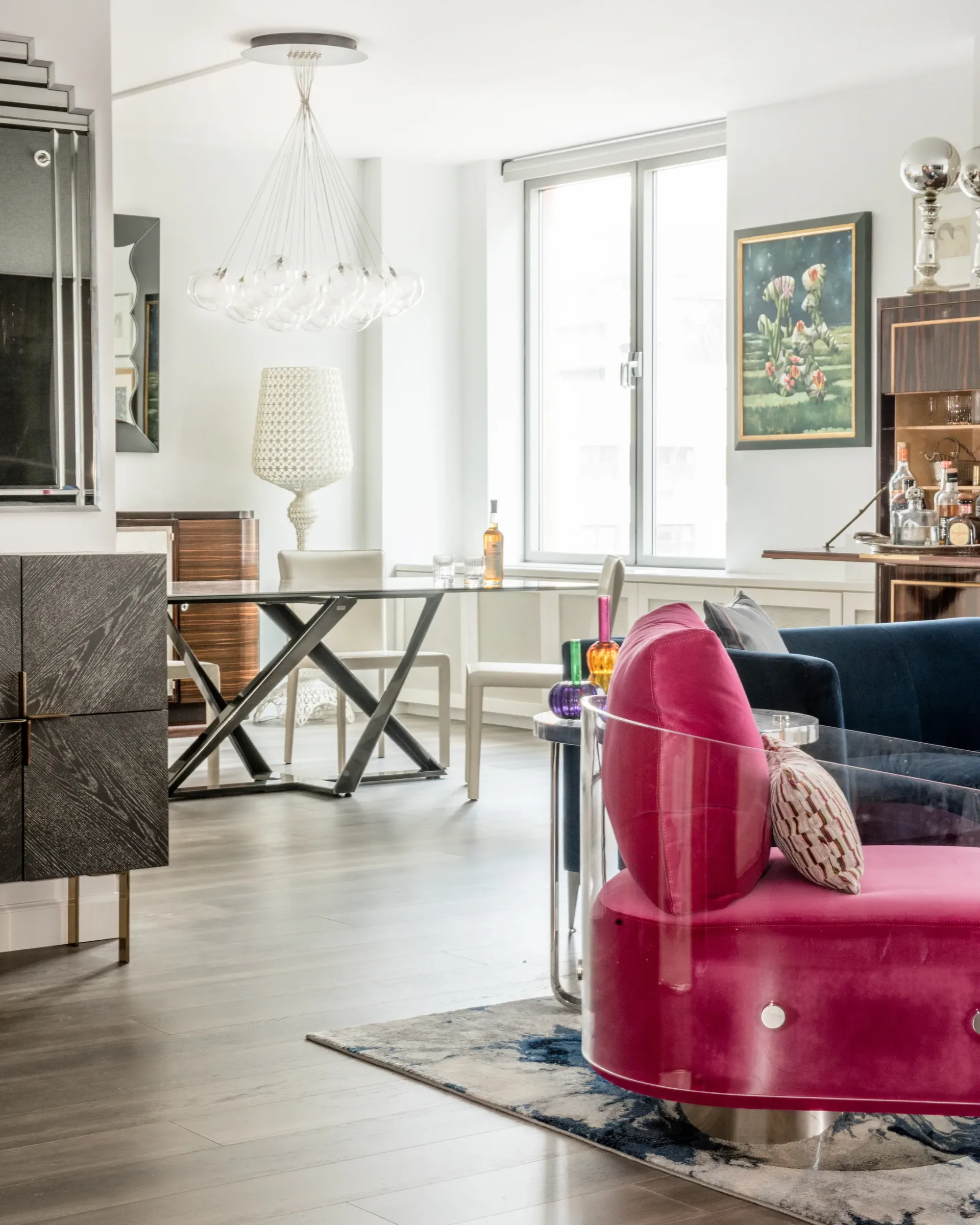 Modern living room featuring a pink velvet curved sofa, wooden desk with X-frame base, pendant lighting, and large windows, designed by Cori Thune.
