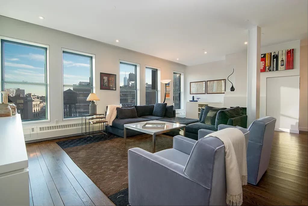 Modern high-rise living room with floor-to-ceiling windows overlooking city skyline, featuring gray upholstered seating, dark wood coffee table, and warm wood flooring by Cori Thune.