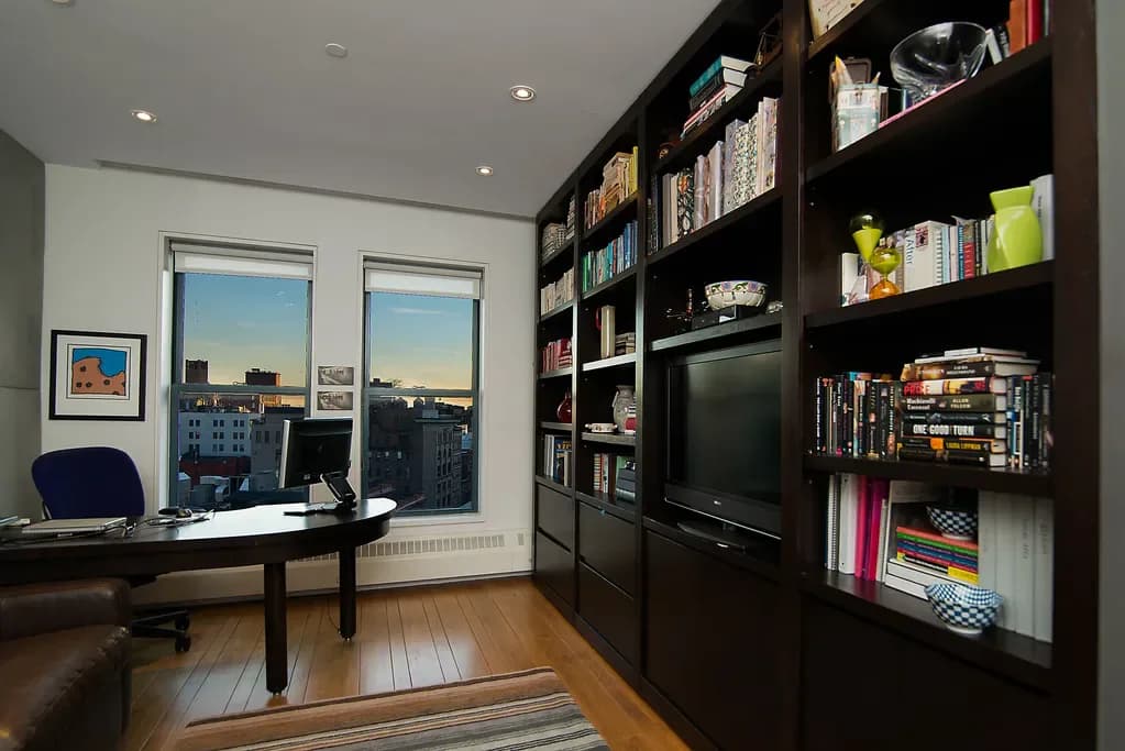 Modern home office featuring dark built-in shelving with TV and storage, black desk by windows overlooking city skyline, wood flooring, and white walls by Cori Thune.