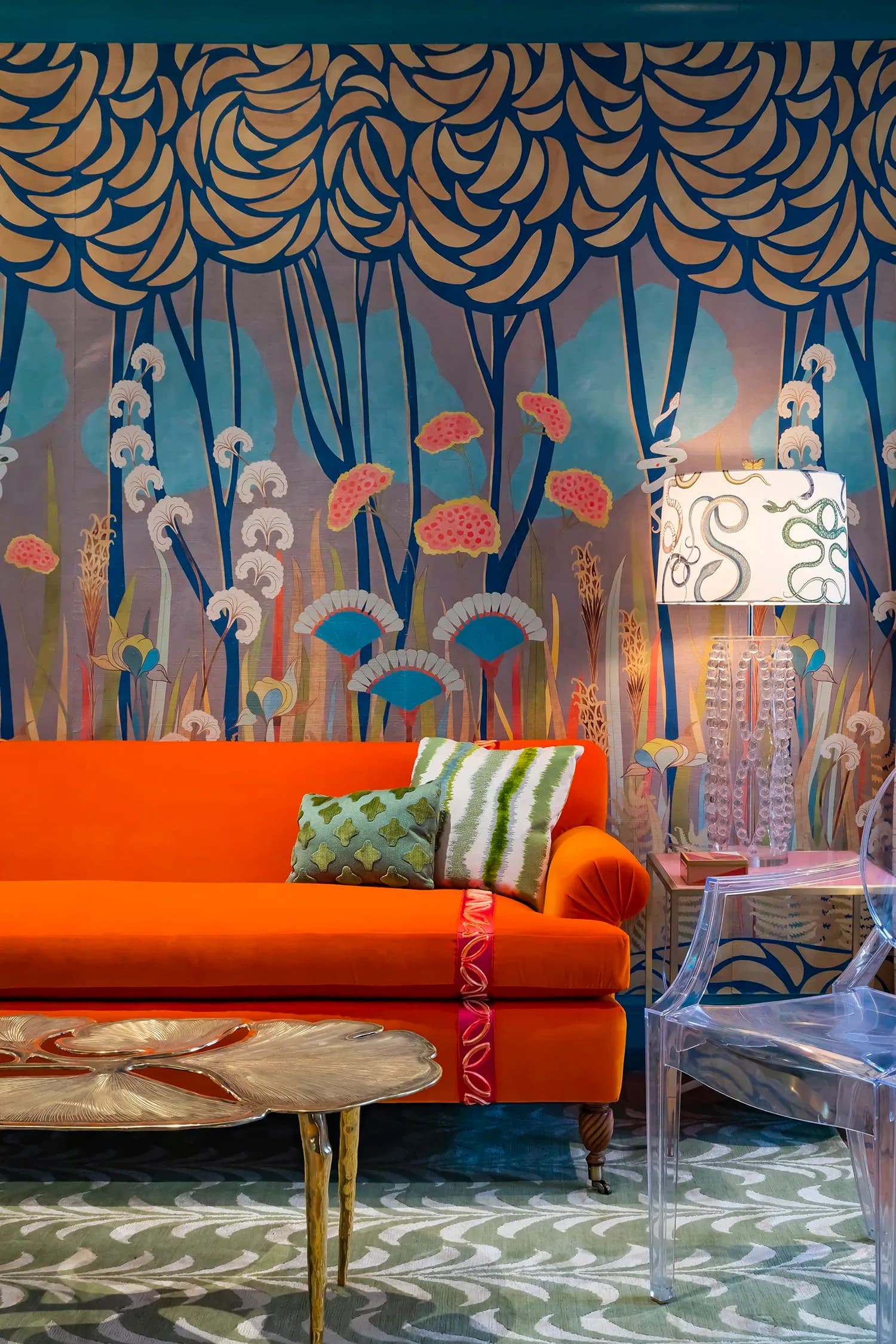 A vibrant interior featuring a bold orange sofa with patterned cushions, positioned against a teal wall mural with stylized botanical and eye motifs in orange, blue, and cream tones, by Courtney McLeod.