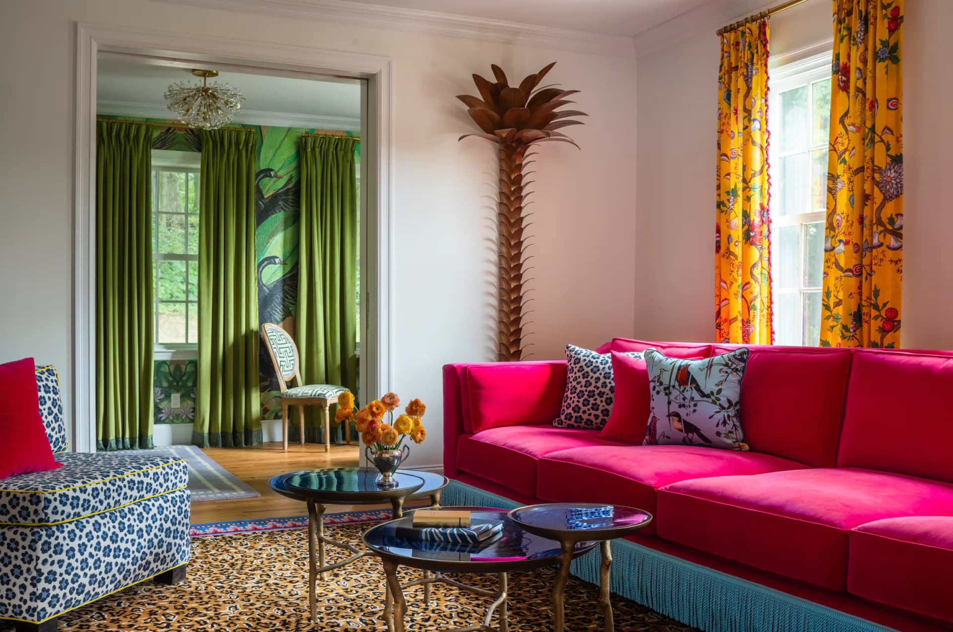 Vibrant living room with hot pink sofa, patterned accent chair, palm tree sculpture, green curtains, and colorful patterned fabrics by Courtney McLeod.