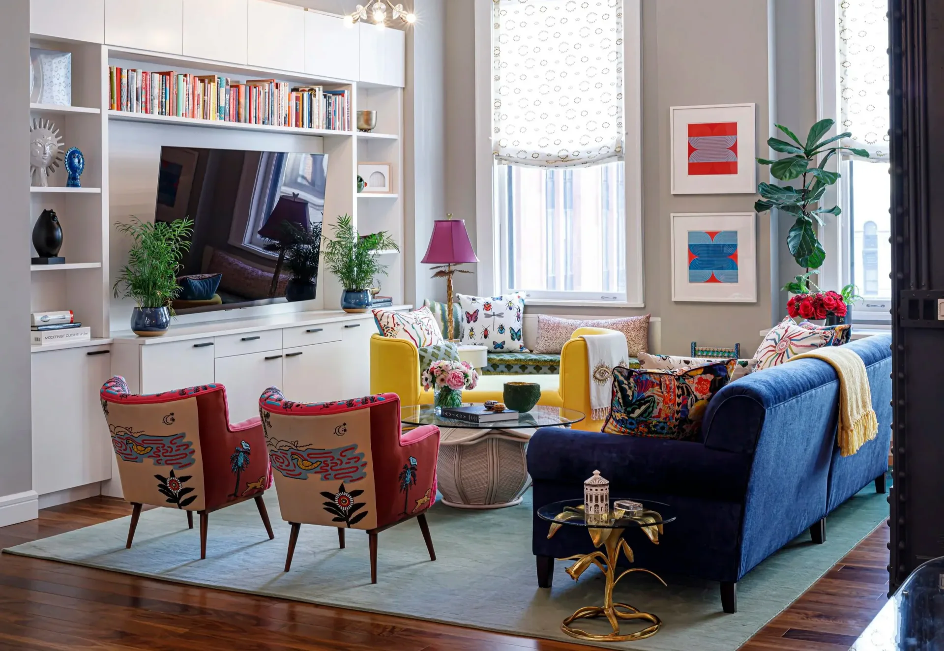 Colorful living room with blue sofa, red accent chairs, built-in white cabinetry, and warm wood floors by Courtney McLeod.