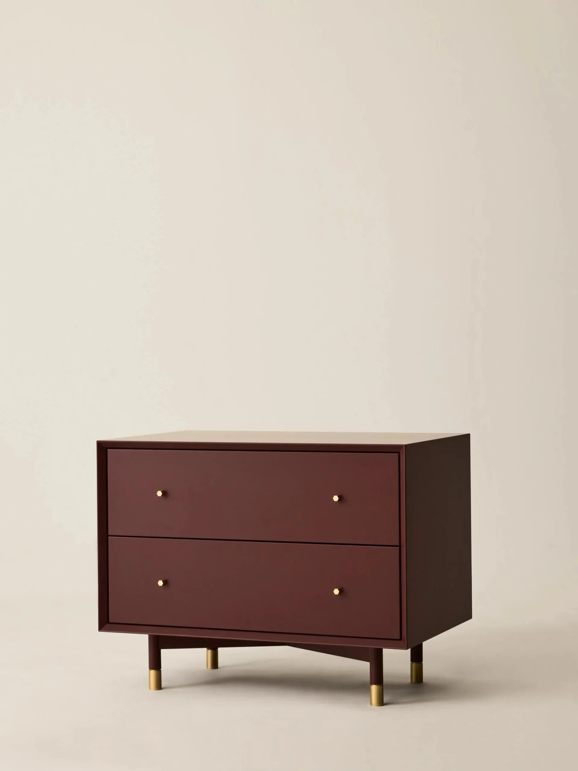A burgundy mid-century modern nightstand with two drawers and brass legs against a neutral beige background by Crystal Ellis.