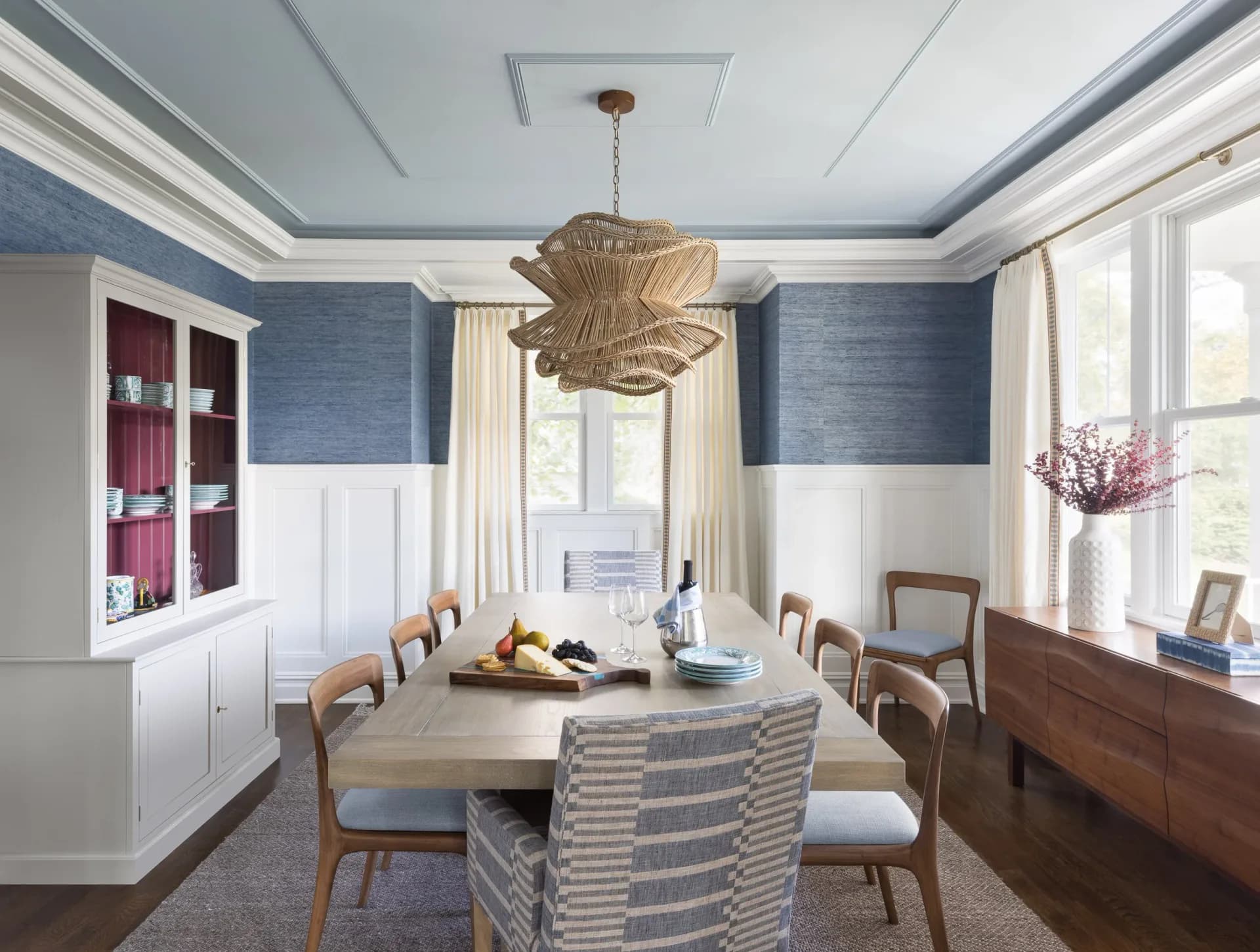 Contemporary dining room with blue textured walls, white wainscoting, natural wood table, striped upholstered chairs, woven pendant light, and cream curtains by Crystal Maki.