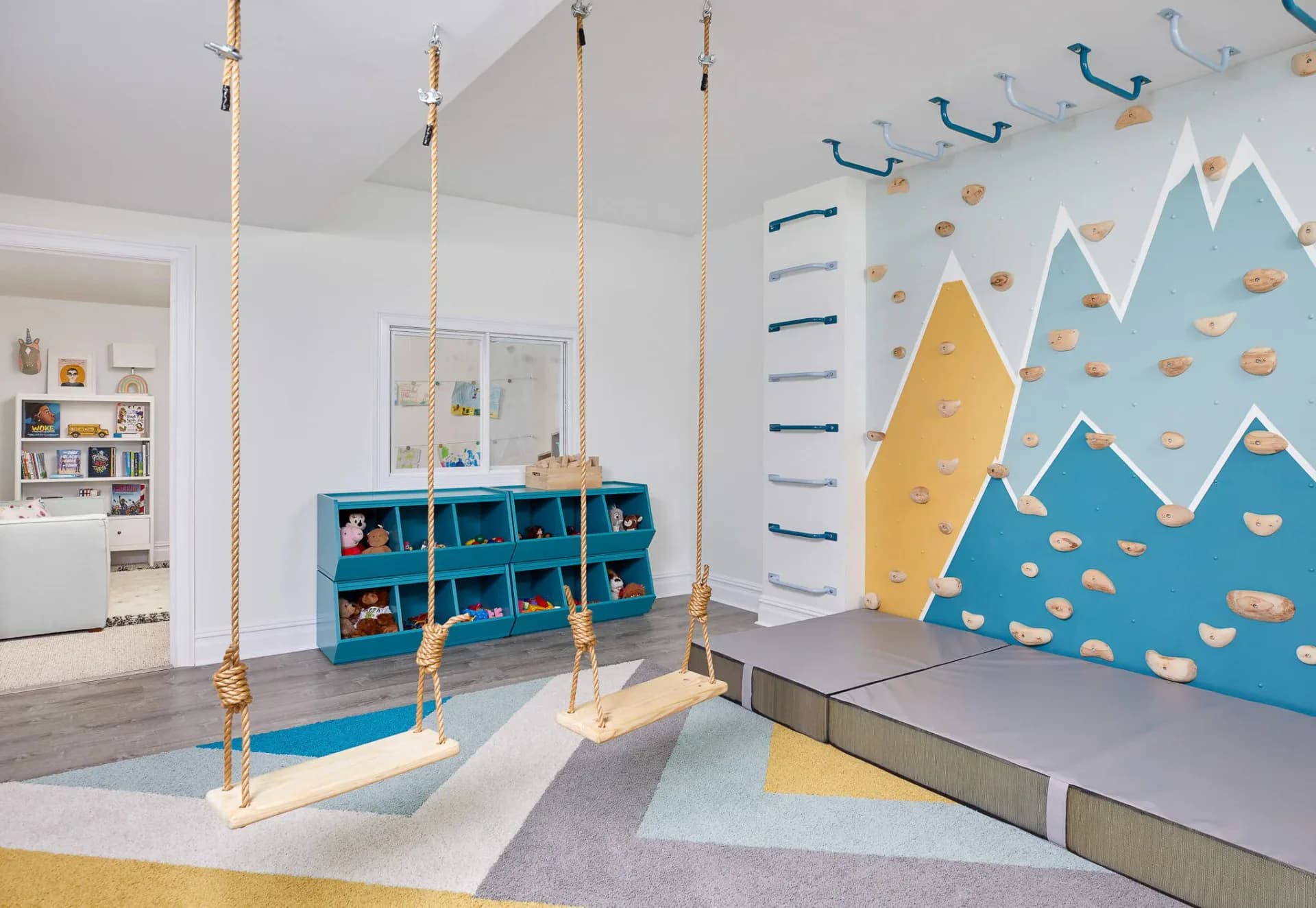 Bright playroom with rope swings, teal storage cubioids, blue mountain wall mural, and cloud details by Crystal Maki.