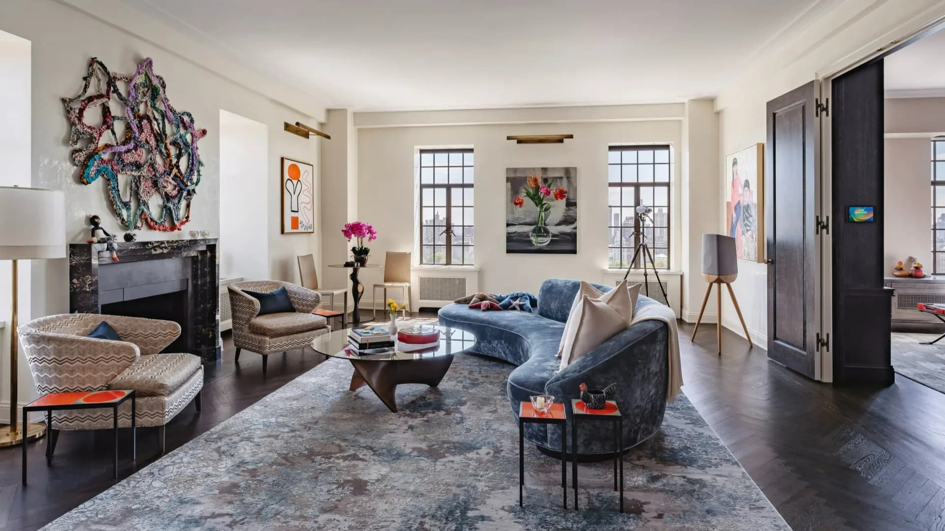 Sophisticated living room with dark hardwood floors, gray area rug, blue velvet sofa, black fireplace with artwork, brass accents, and modern furnishings by Cynthia Peralta-Brito.