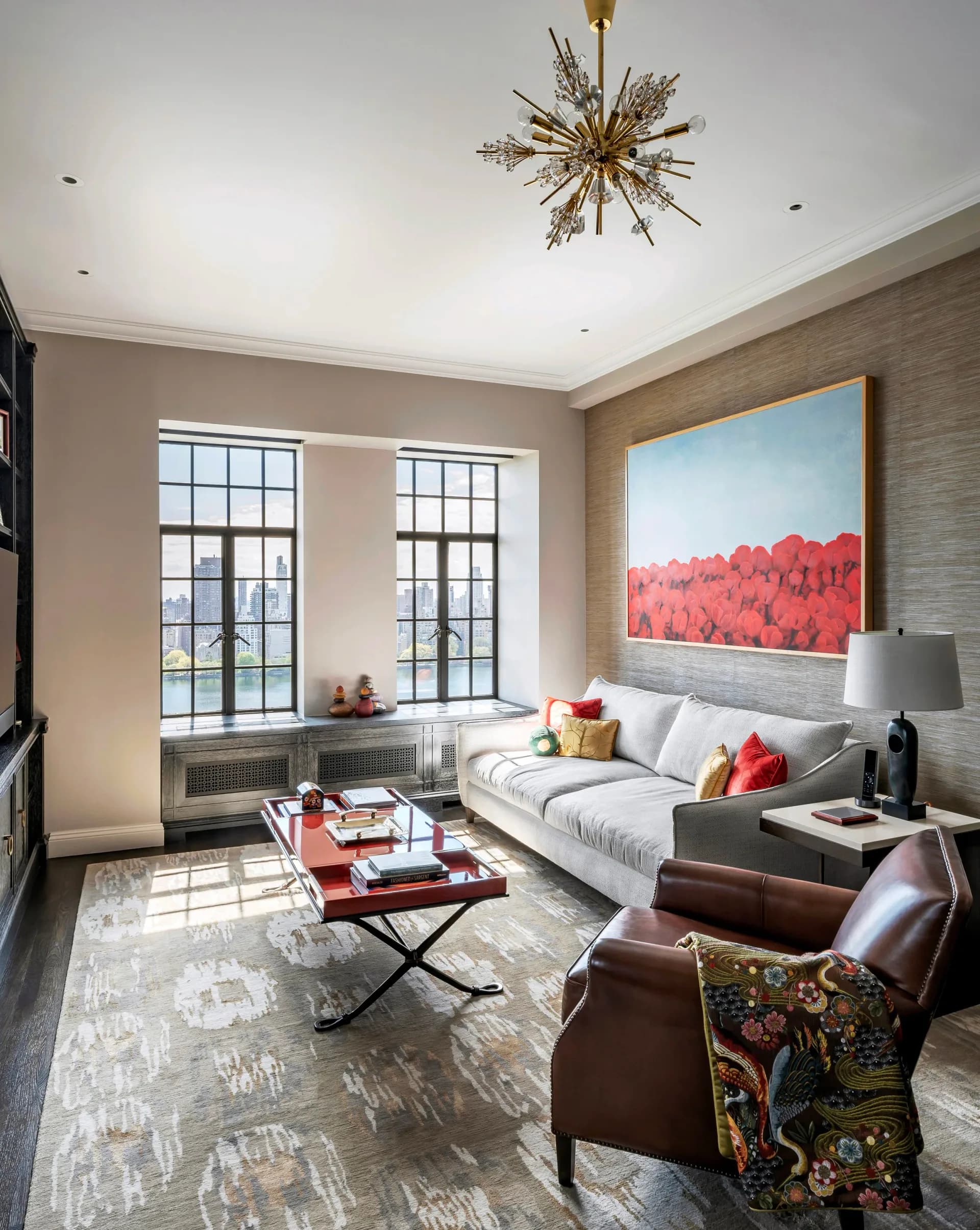A contemporary living room with taupe textured walls, large gridded windows, a gray upholstered sofa with coral pillows, leather accent chairs, a brass starburst chandelier, and a red-pink landscape artwork, designed by Cynthia Peralta-Brito.