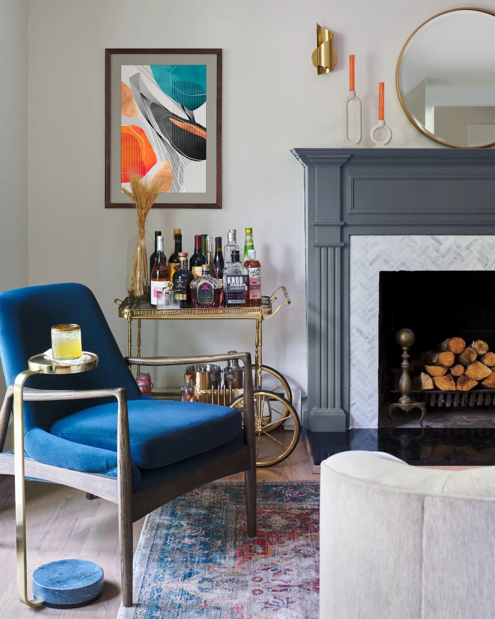 Contemporary living room with blue velvet chair, brass bar cart, dark fireplace, and geometric abstract artwork by Cynthia Peralta-Brito.