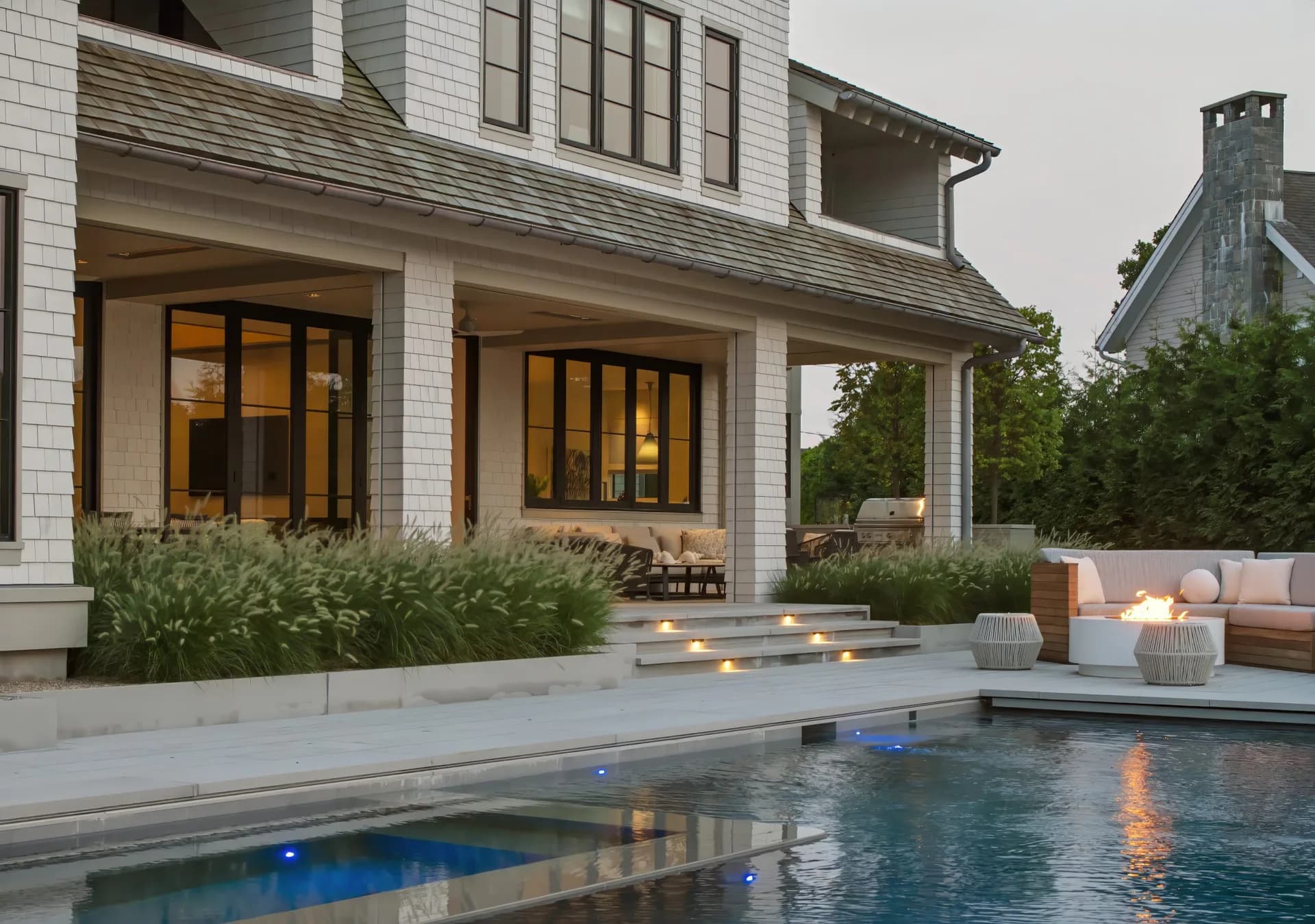 Modern coastal home exterior with white shiplap, large glass doors, and pool with blue lighting at dusk by Cynthia Vengrow.