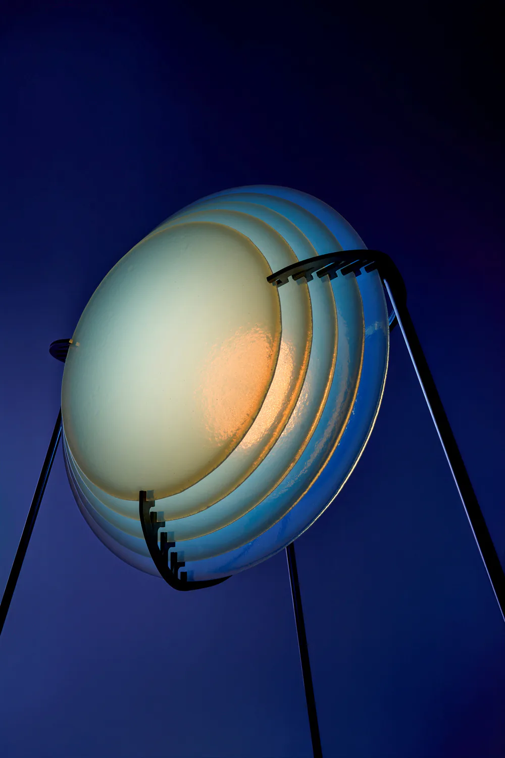 Sculptural spherical lighting installation with concentric metal rings in blue and warm tones, mounted on tripod legs against twilight sky by Dace Sūna.