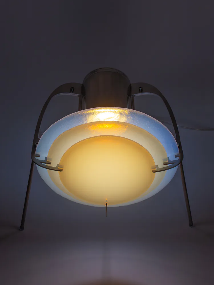 Sculptural pendant lighting fixture with layered dome design in cream and blue tones, suspended from dark metal frame, glowing warmly against dark background by Dace Sūna.