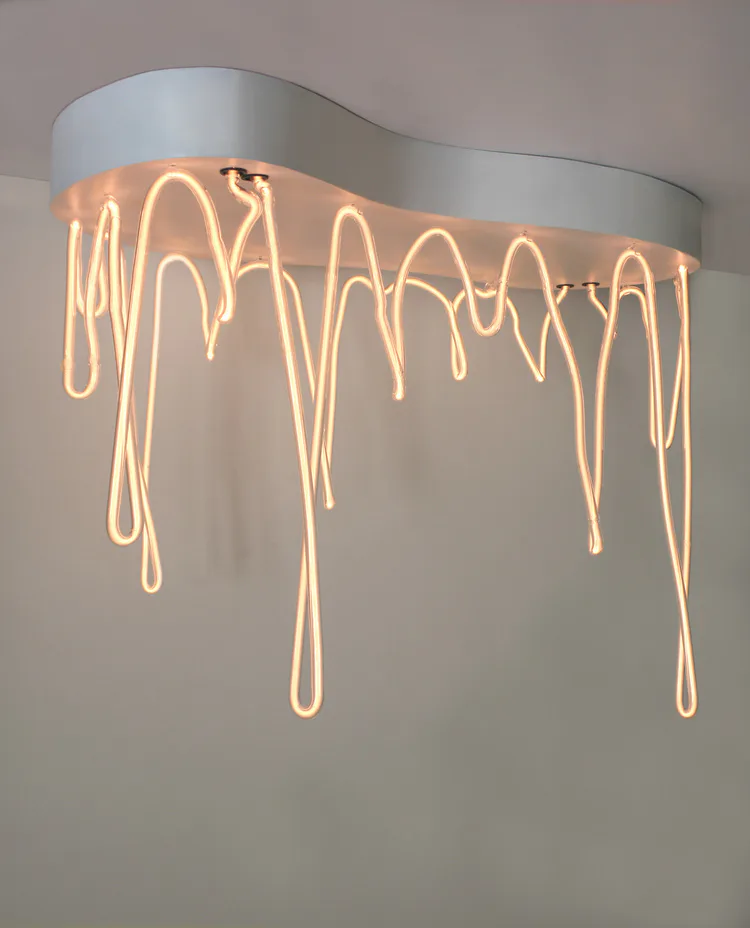 Sculptural ceiling installation featuring glowing neon tubing cascading downward from a curved gray plaster base, creating an organic draping effect with warm amber light by Dace Sūna.