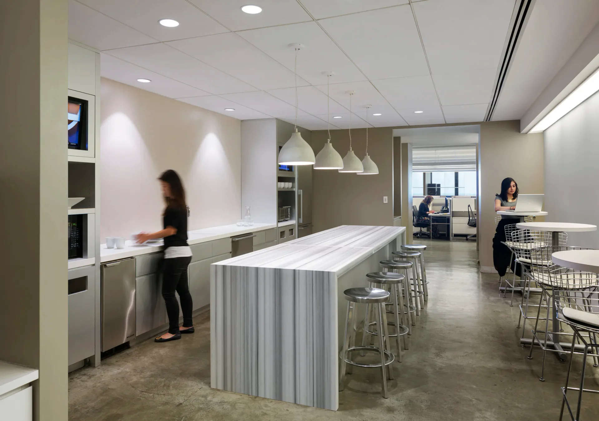 Modern office break room with central white striped island counter, pendant lighting, concrete floors, and neutral palette by Damiano Geraci.