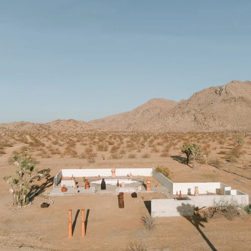 Desert landscape installation with white architectural elements and orange sculptural pieces arranged on sandy terrain with sparse vegetation, by Dan John Anderson.