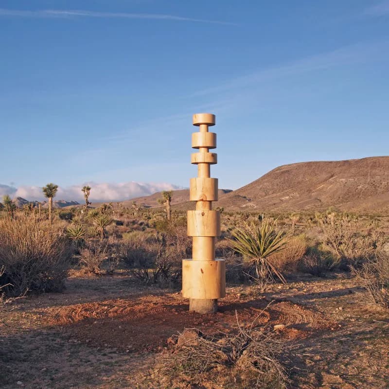 Desert landscape installation featuring a tall stacked wooden sculptural tower amid arid terrain with scattered vegetation and mountains beyond, by Dan John Anderson.