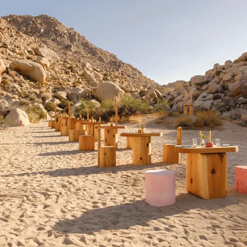Desert landscape installation with wooden sculptural elements and colored cylindrical forms arranged on sand, surrounded by rock formations, by Dan John Anderson.