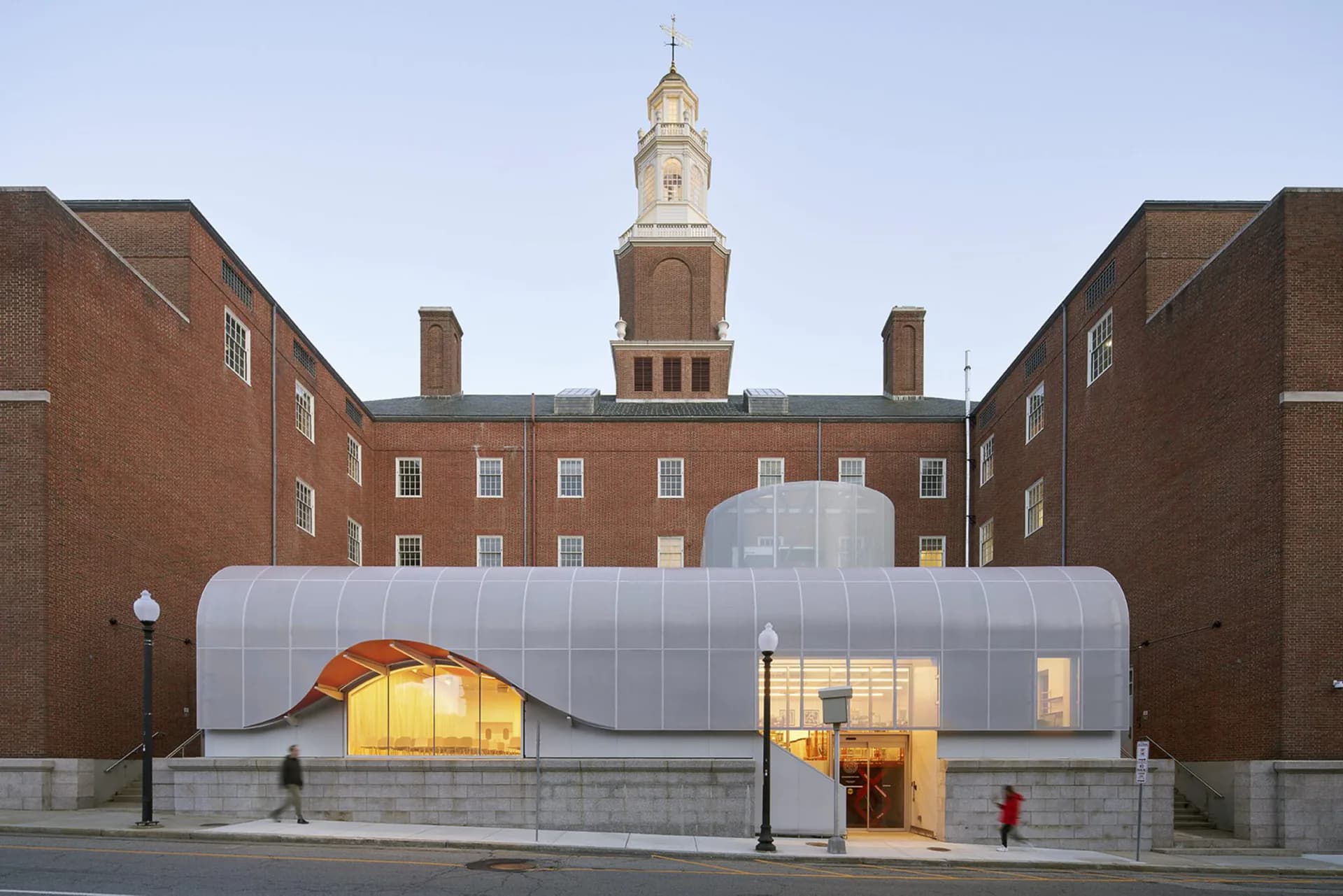 Translucent curved pavilion with glowing interior nestled between historic brick buildings, featuring undulating metallic facade and illuminated entrance by Dan Wood.