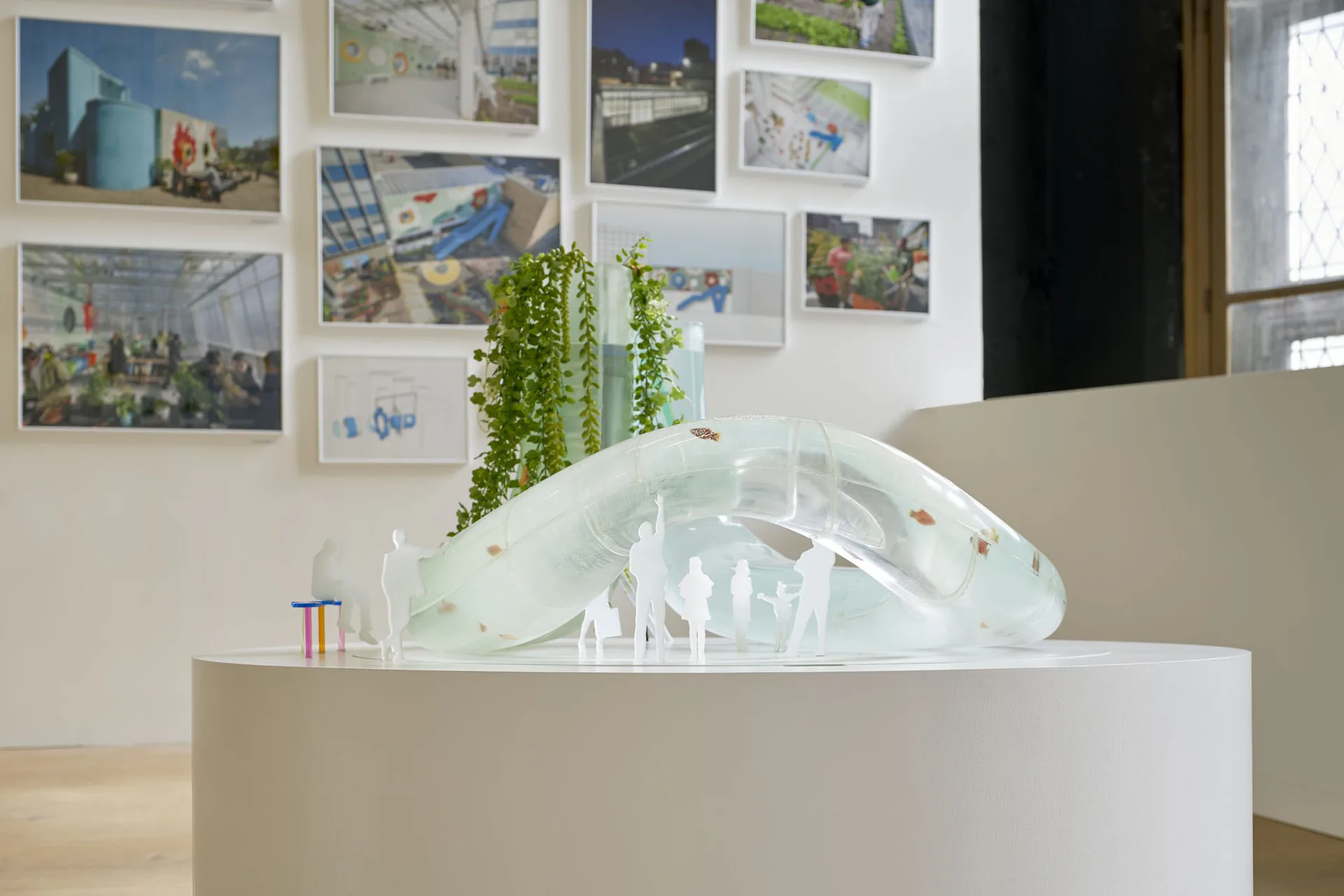 White gallery space with sculptural architectural model on pedestal, hanging plants, and travel photography gallery wall installation by Dan Wood.