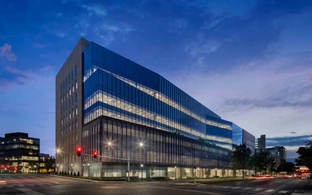 Modern glass-clad office building with horizontal banding lit from within at dusk, featuring a striking curved facade and ground-level glazing by Dana McCarty.