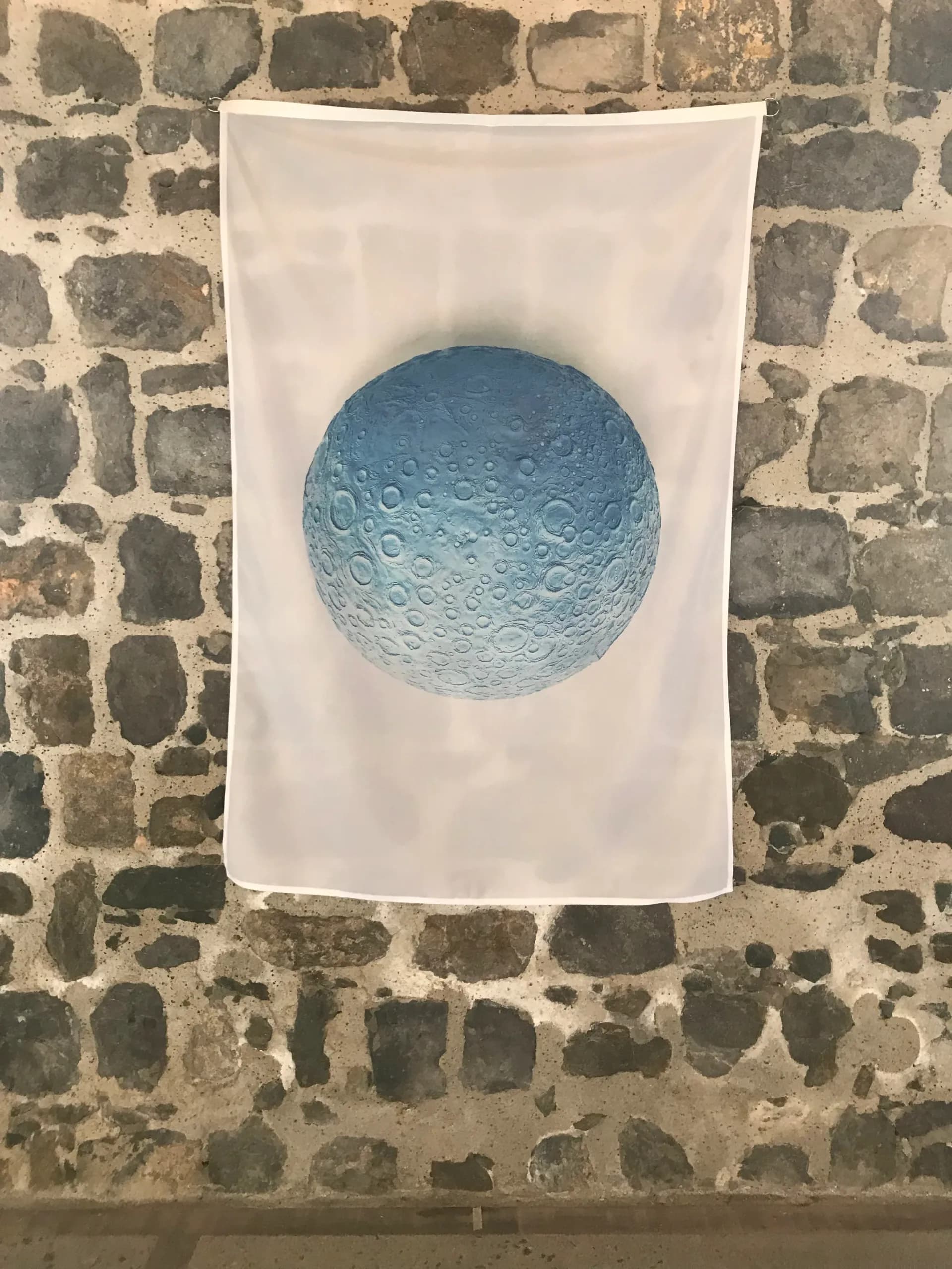 Blue moon sculpture with crater details mounted on white fabric against weathered stone wall by Daniel Arsham.