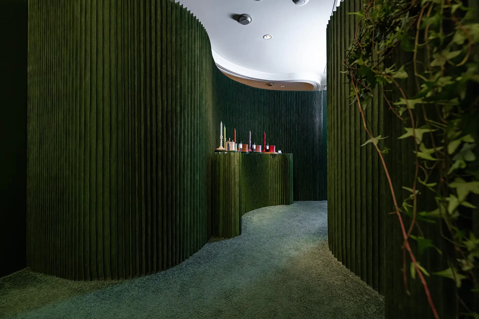 A curved green corrugated corridor with skylights opens into a gallery space featuring illuminated candles on a shelf, designed by Daniel Maldonado.