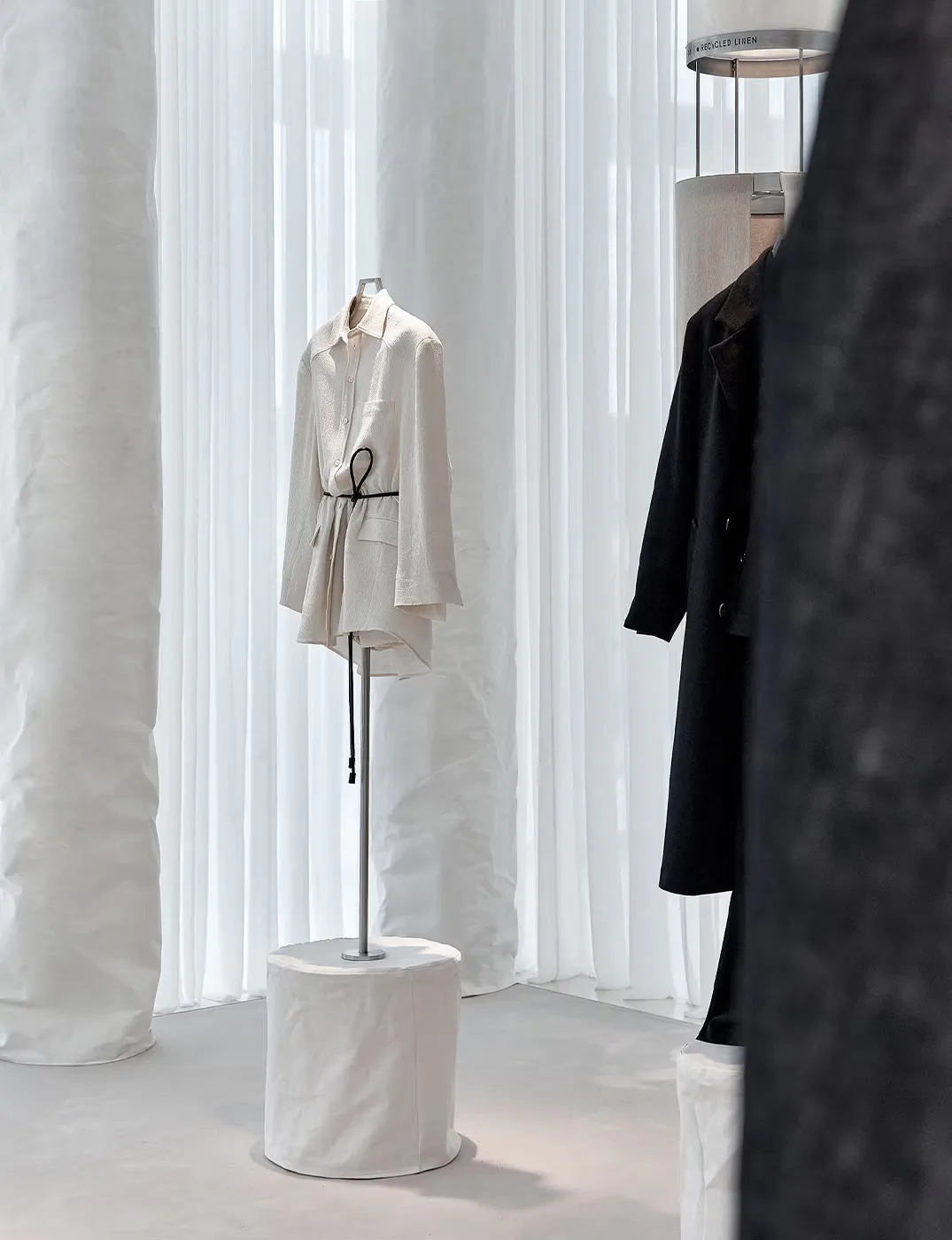 Minimalist clothing display with a beige linen coat on a pedestal amid white curtains and a black garment, designed by Daniel Maldonado.