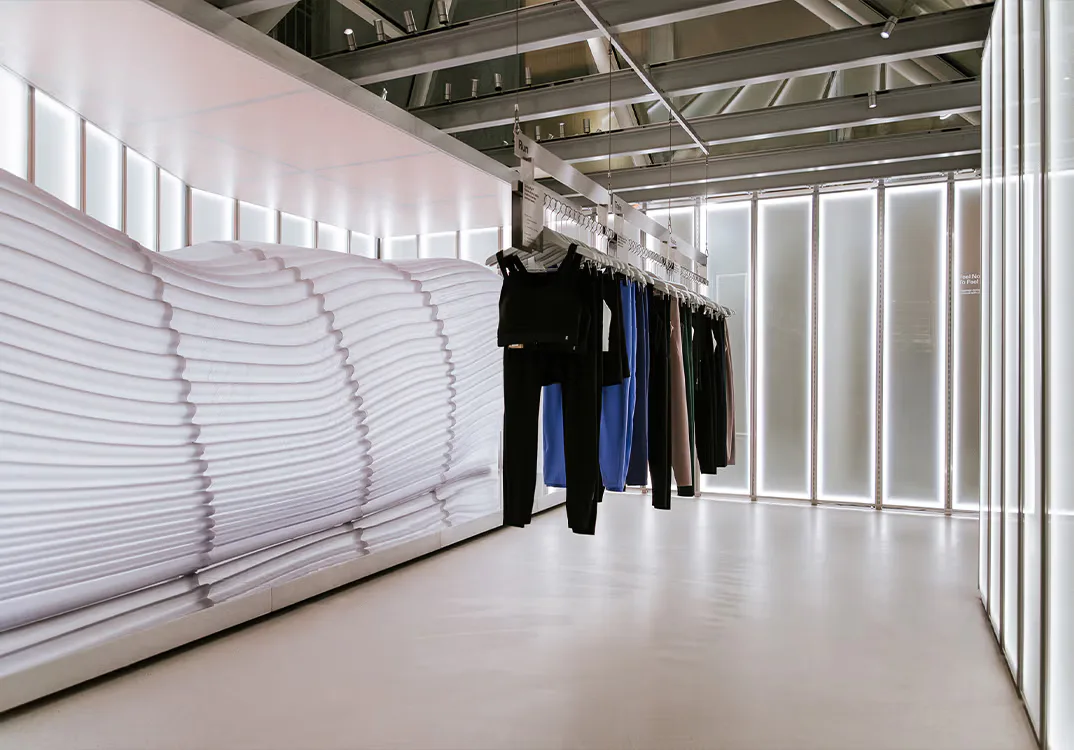 Minimalist retail space with white corrugated wall paneling, polished concrete floor, and hanging black and blue garments, designed by Daniel Maldonado.