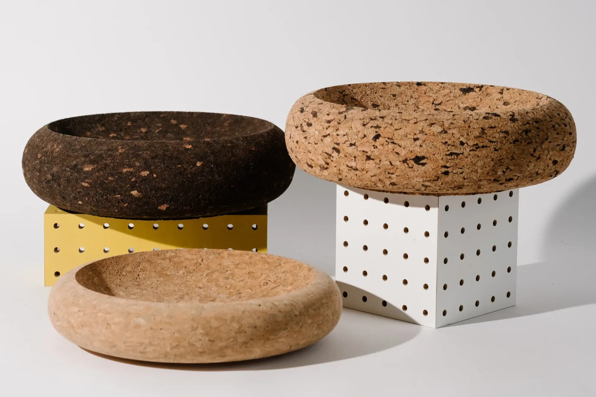 Three cork sculptural vessels with organic forms in varying sizes and finishes, displayed on geometric bases in yellow, white, and natural tones by Daniel Michalik.