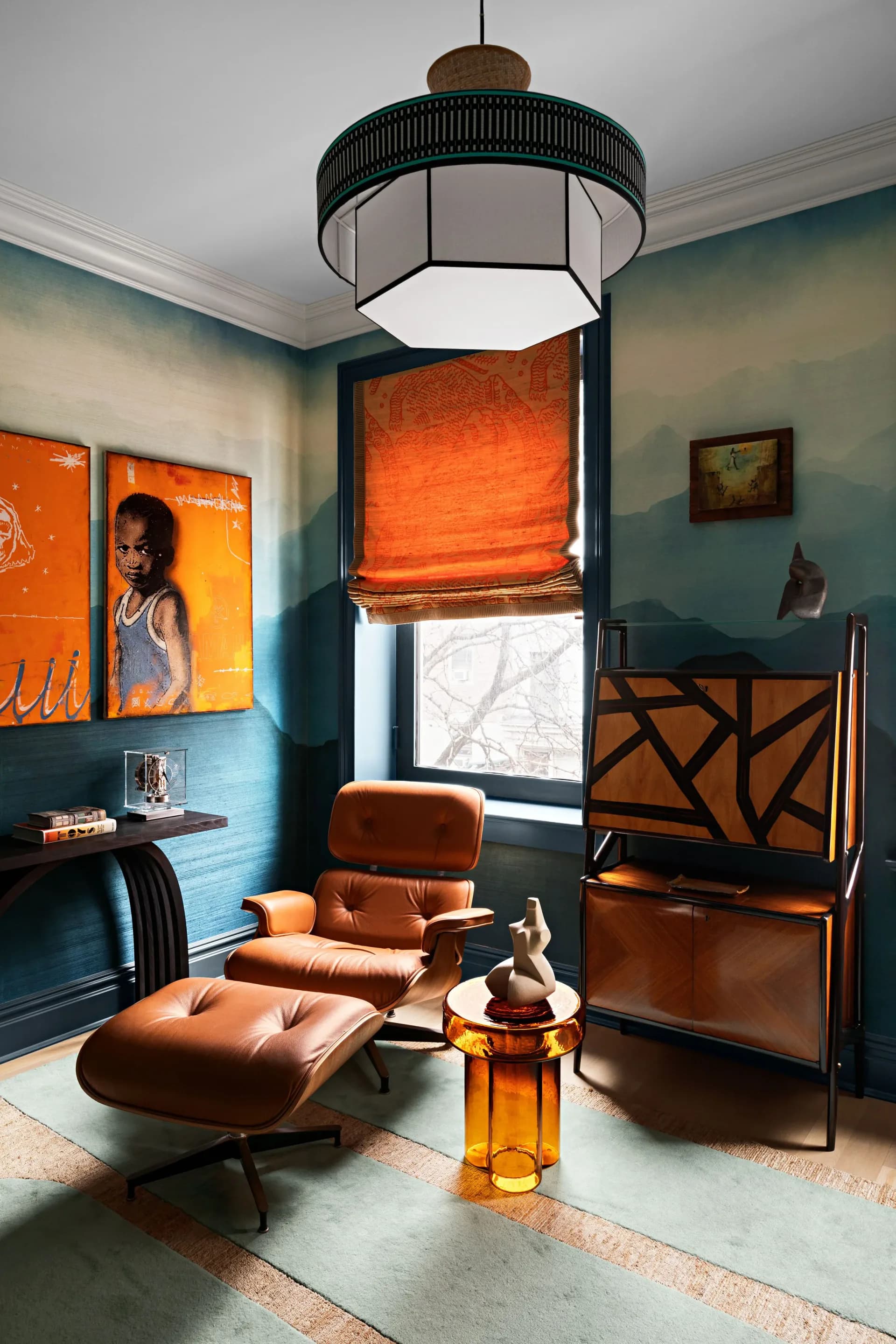Teal-walled den with cognac leather lounge chair, orange roman shade, geometric cabinet, and pendant lighting by Danielle Colding.