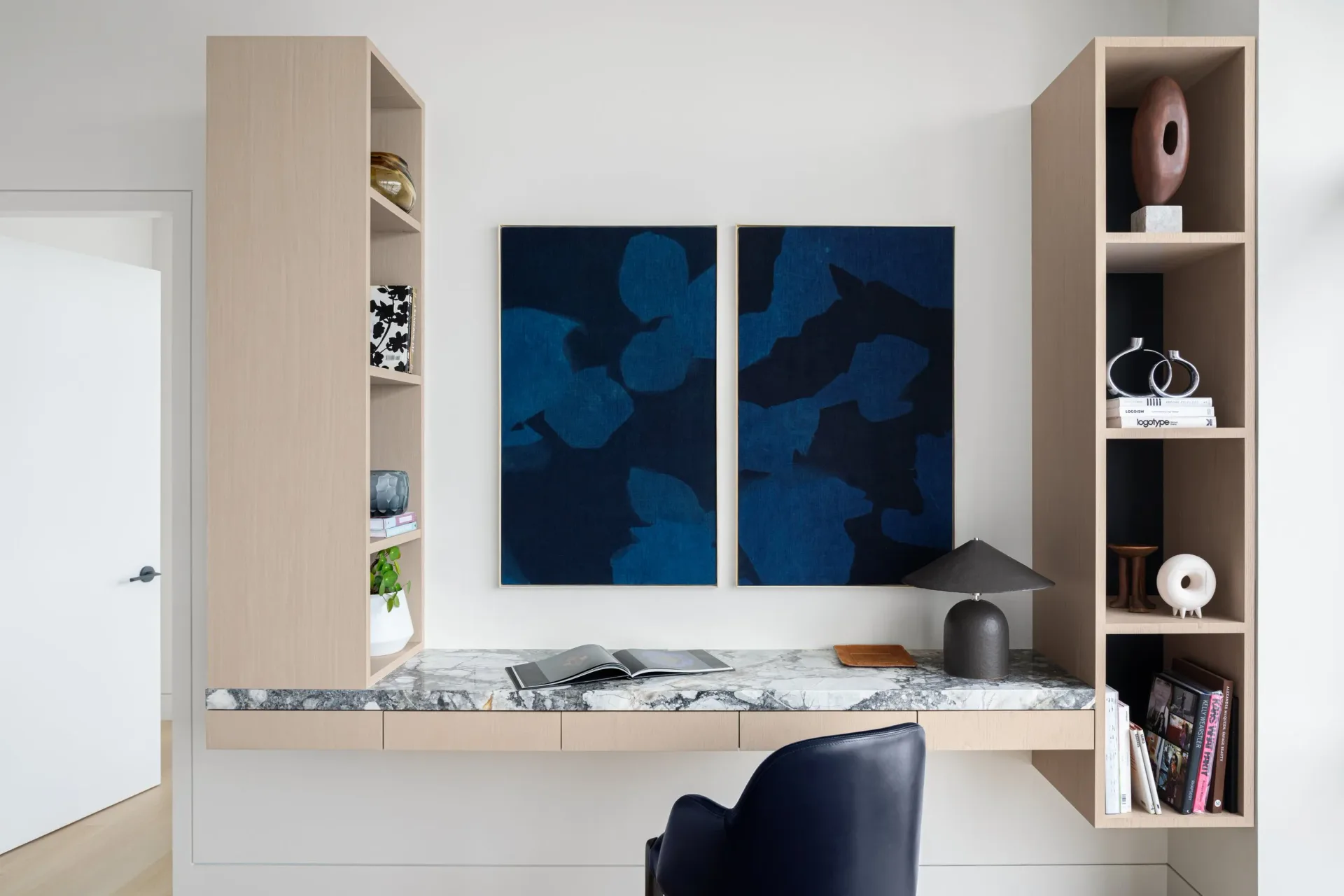 Modern bedroom workspace with built-in shelving, marble desk, navy abstract artwork, and light wood cabinetry by Danielle Colding.