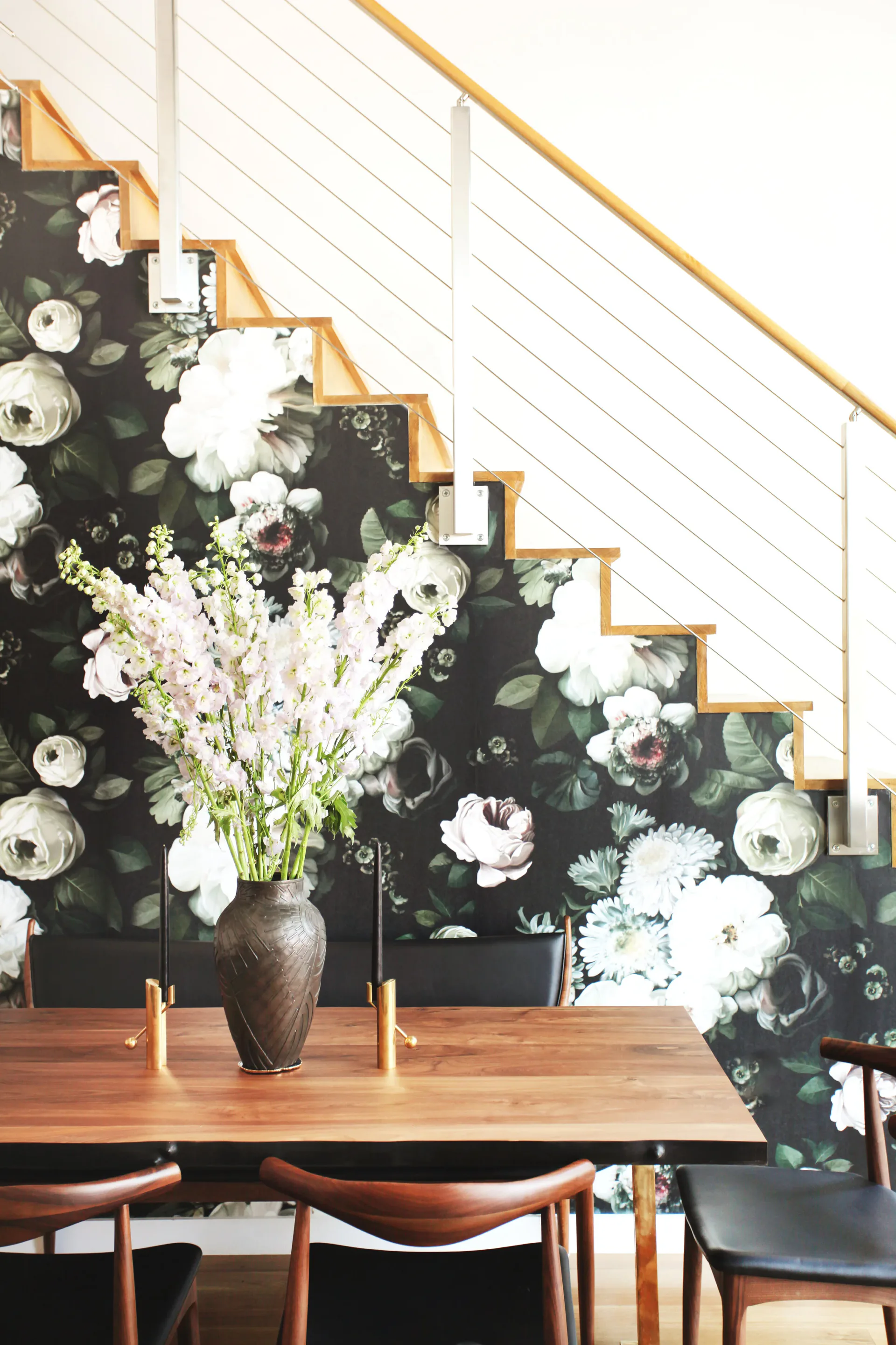 Dining space with wooden table and mid-century chairs beneath a staircase, featuring a dark botanical wallpaper and fresh white flower arrangement by Danielle Colding.