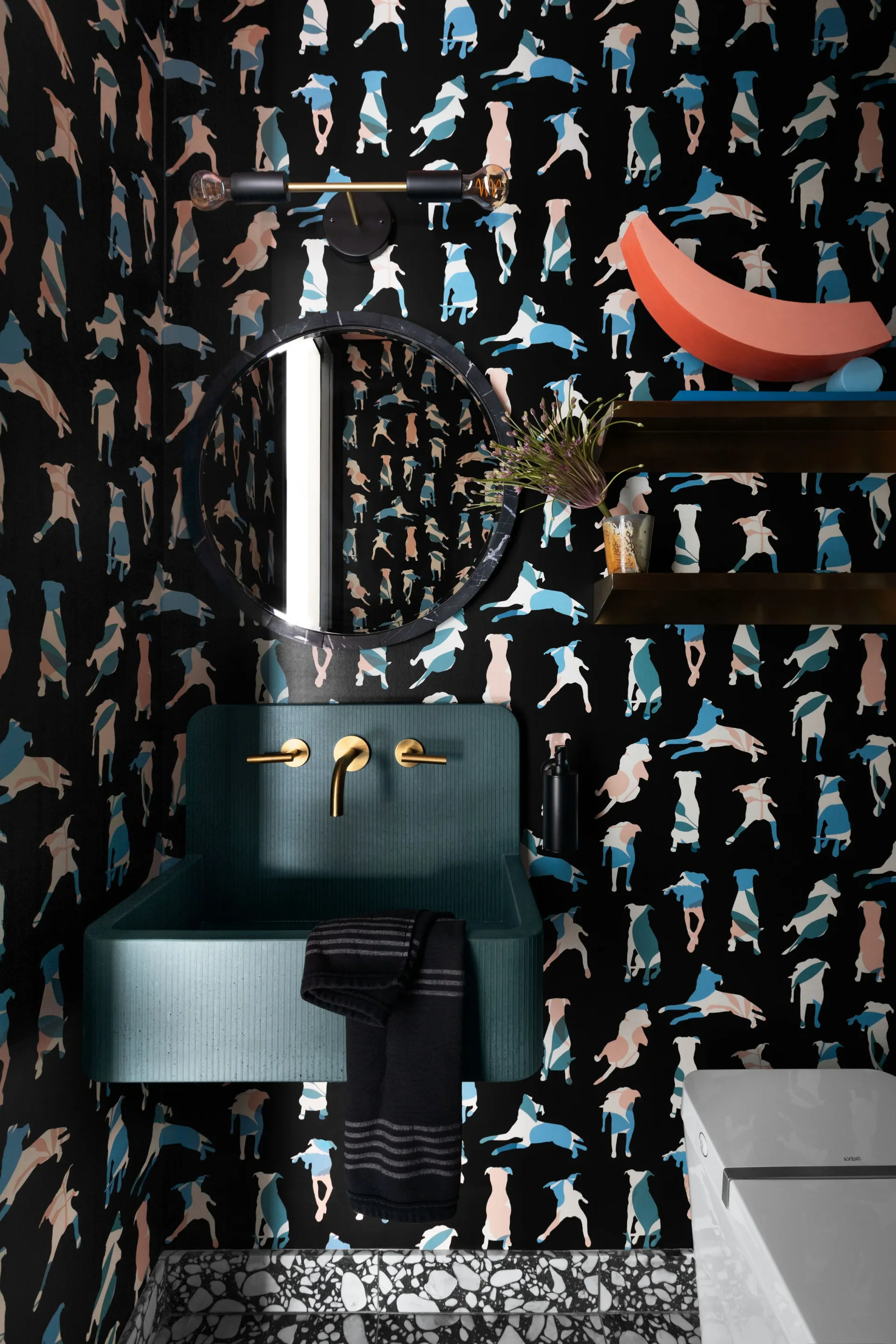 Bold bathroom with black wallpaper featuring illustrated figures in blue and brown tones, teal pedestal sink with brass fixtures, and coral sculptural element by Danielle Colding.