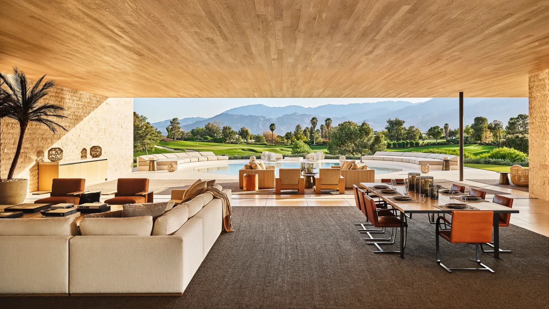 Luxurious open-plan lounge with cream seating, orange accents, and polished wood ceiling overlooking a golf course and mountains by Danielle Peterfy.